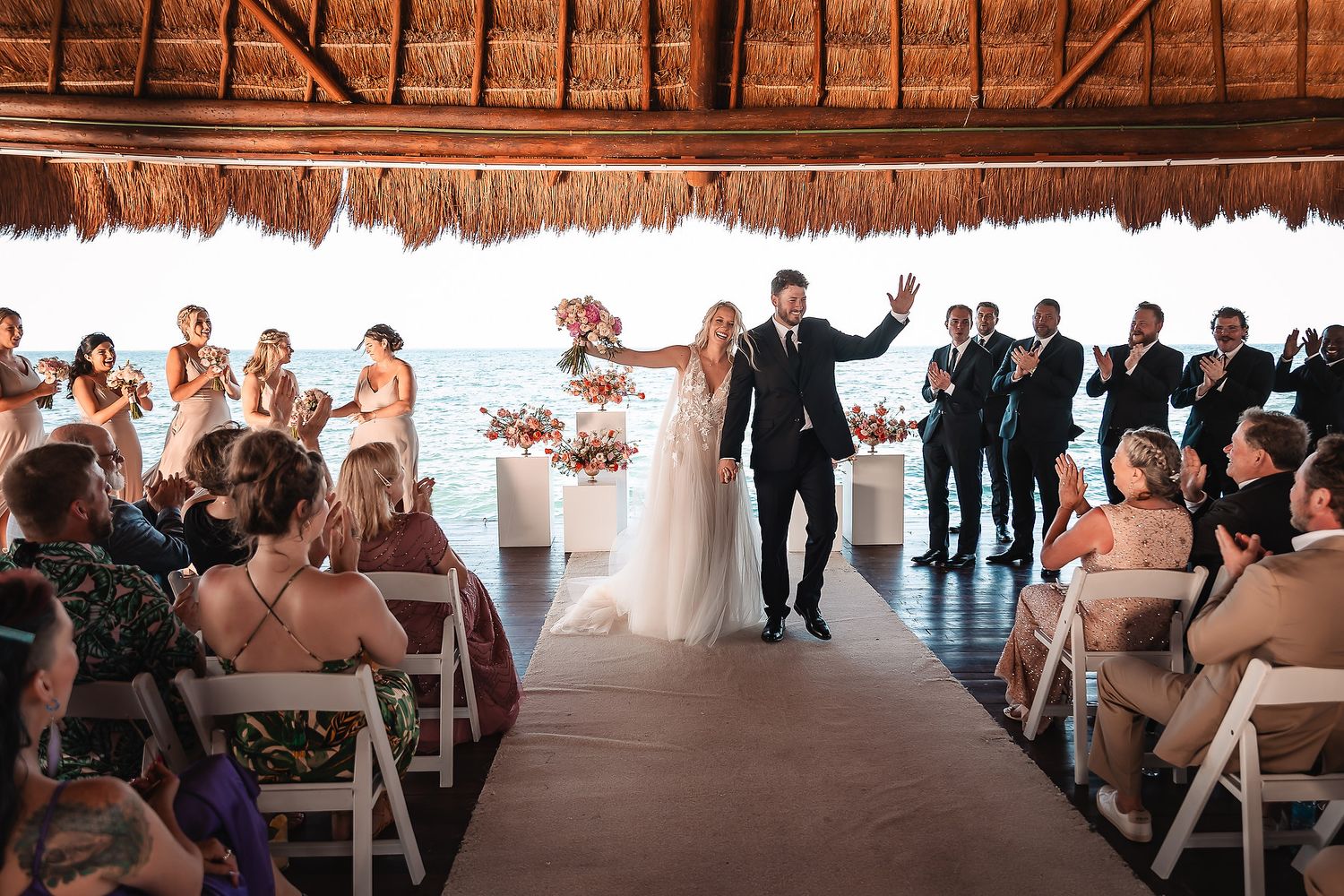 Guests seated in white chairs under a tropical palapa during a destination wedding ceremony.