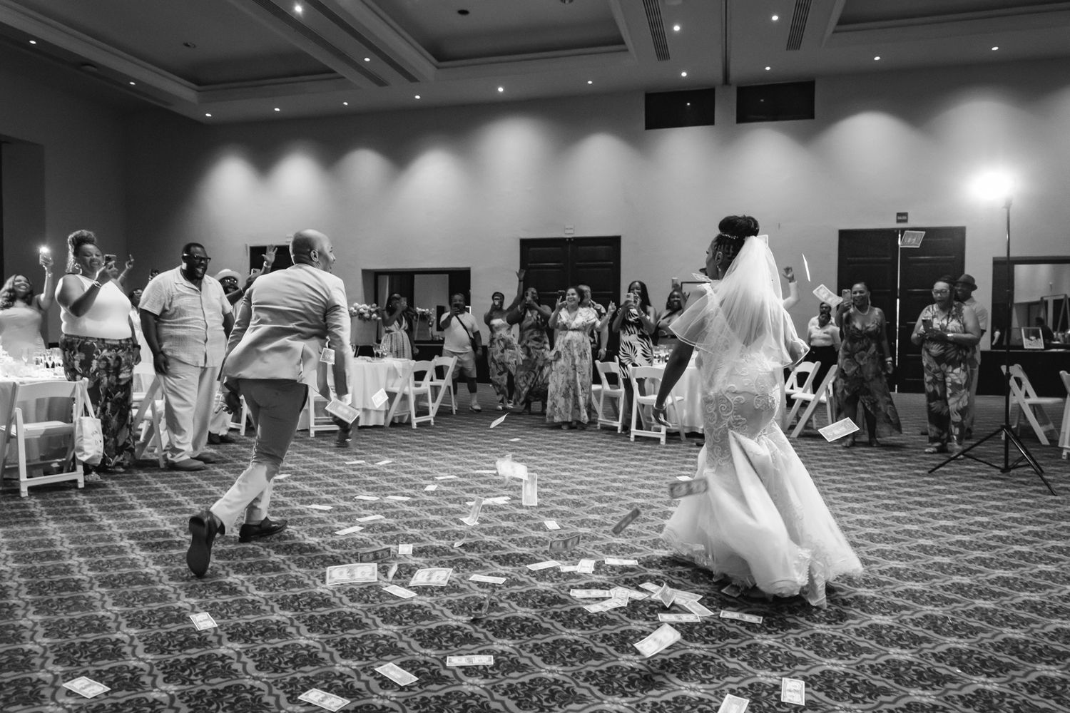 Black and white photo of a bride dancing with guests at a wedding reception, money scattered on the ballroom floor.