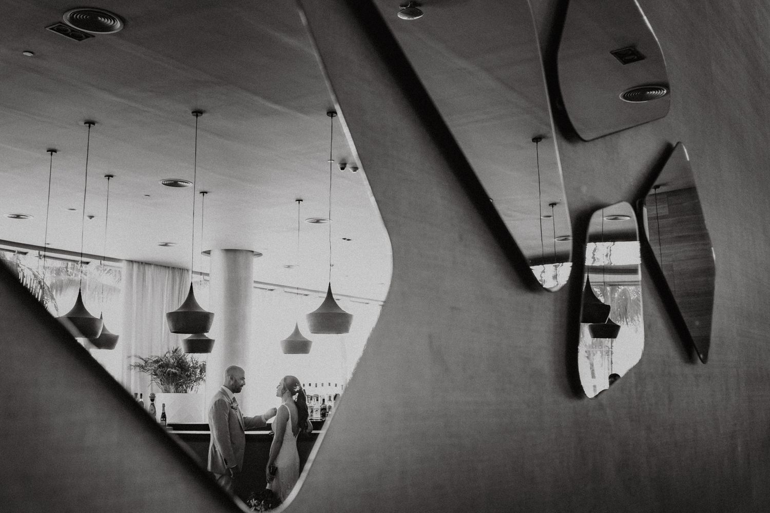 Black and white photo of a person reflected in geometric mirrors on a wall, with pendant lights visible in the reflection.