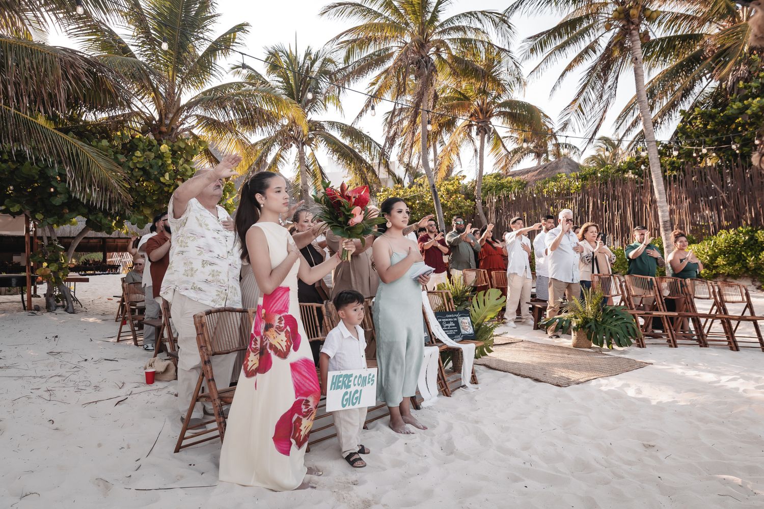 Guests celebrate at a tropical beach wedding ceremony surrounded by palm trees and white sand.