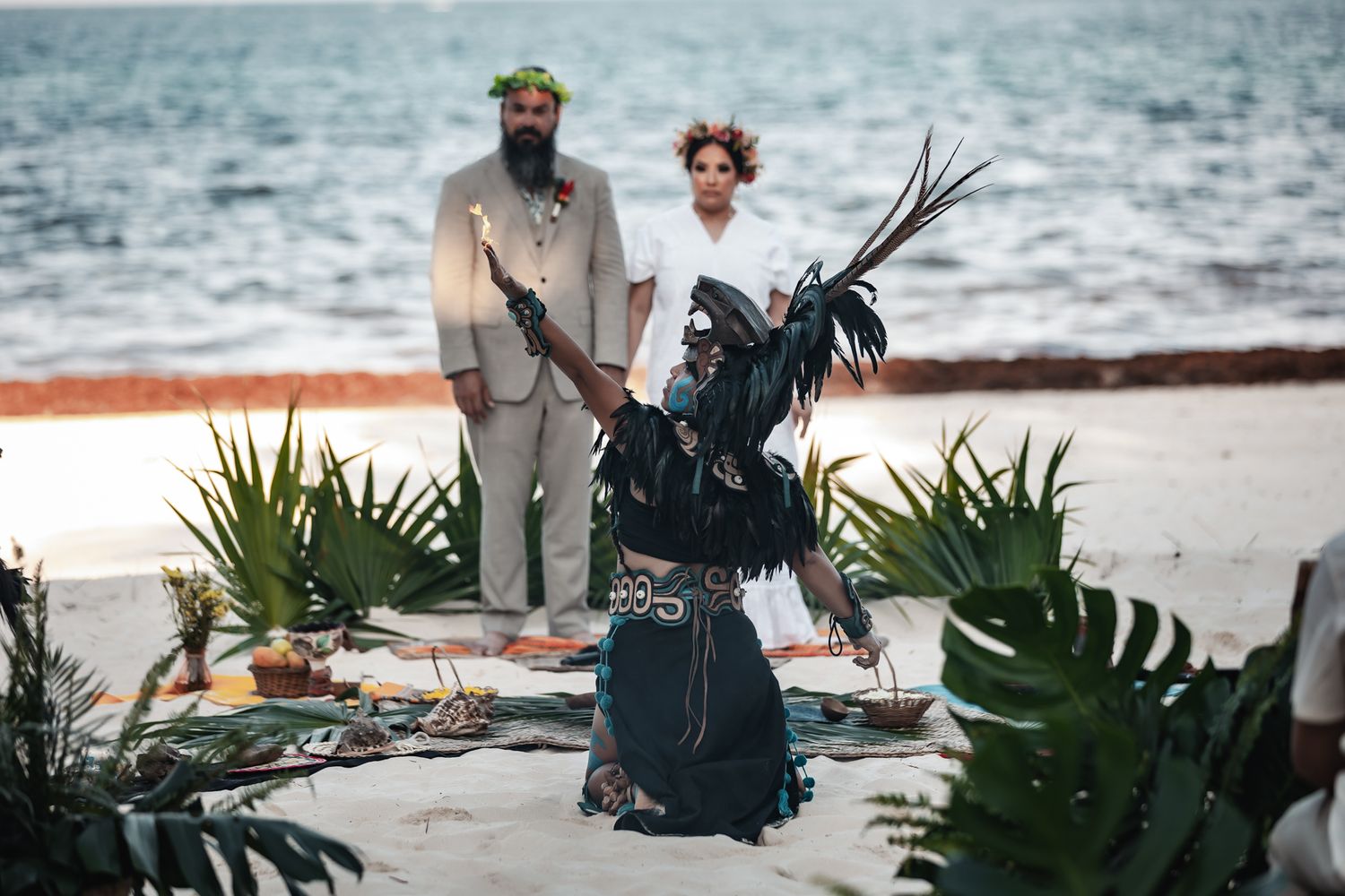 Couple stands at beach wedding ceremony with performer in black feathered costume on sandy shore.