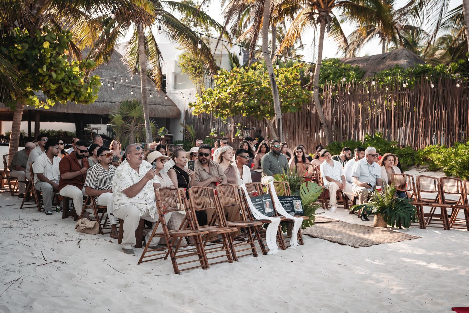 Guests seated on wooden chairs in the sand at an outdoor beach wedding ceremony surrounded by palm trees.