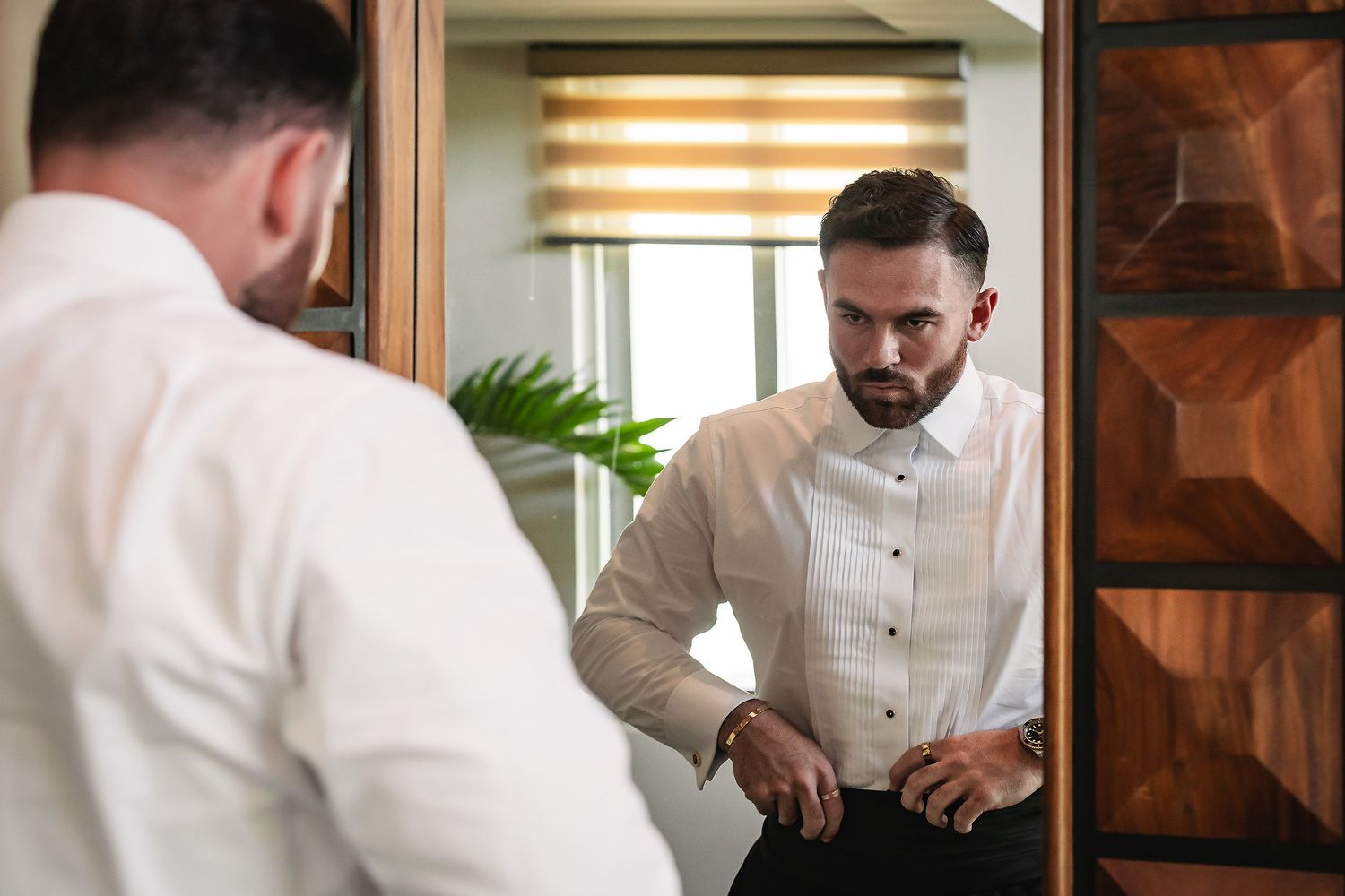 Man in white dress shirt adjusting his belt while looking at his reflection in a mirror.