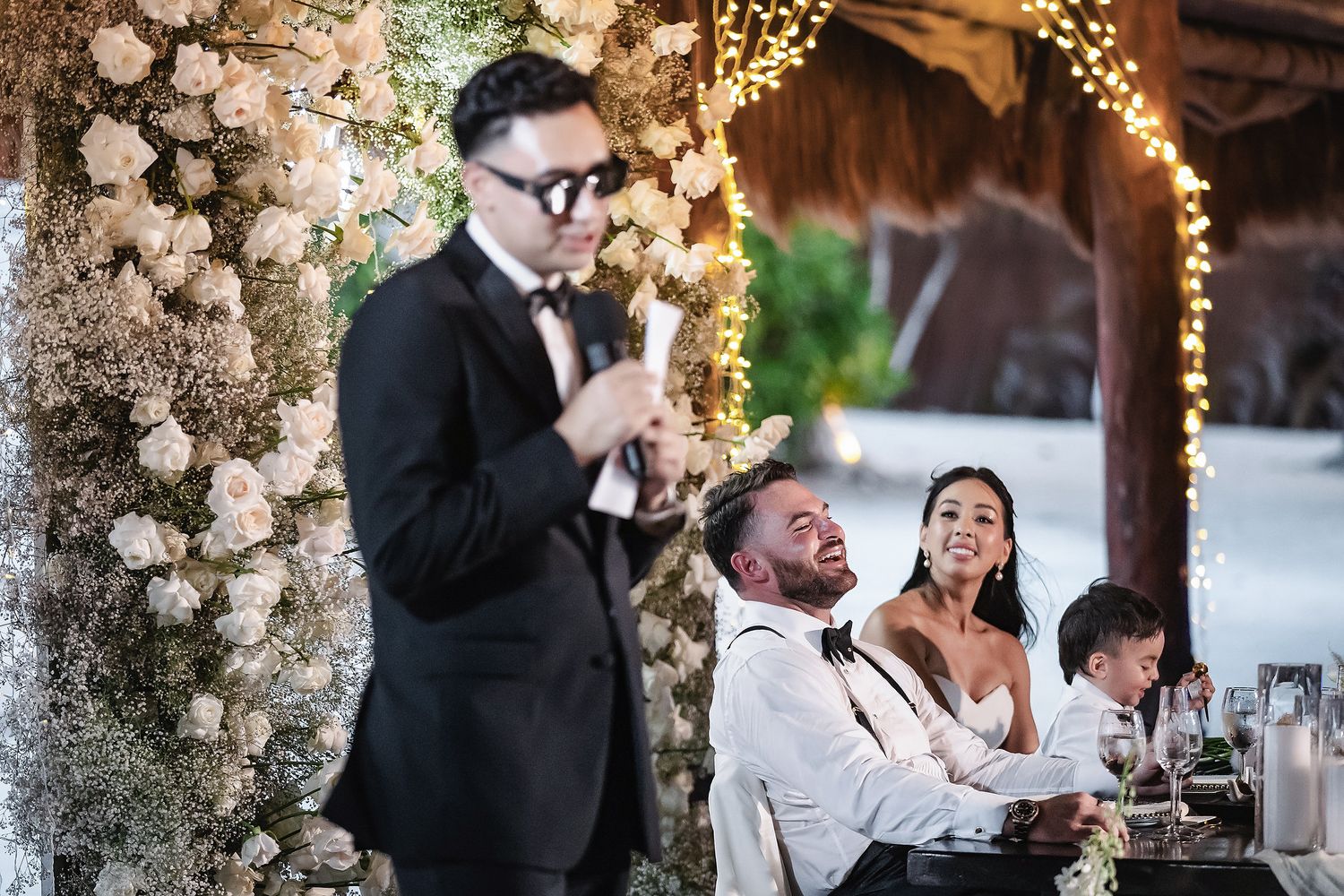 Man in tuxedo giving wedding speech while bride and groom laugh joyfully at floral-decorated reception.