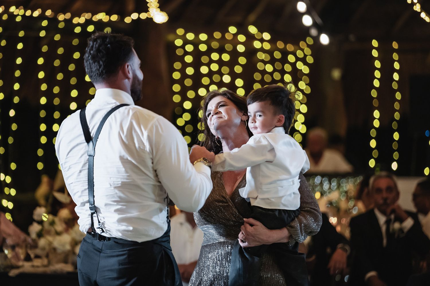 Couple dancing with young boy at elegant wedding reception with warm bokeh string lights in background.