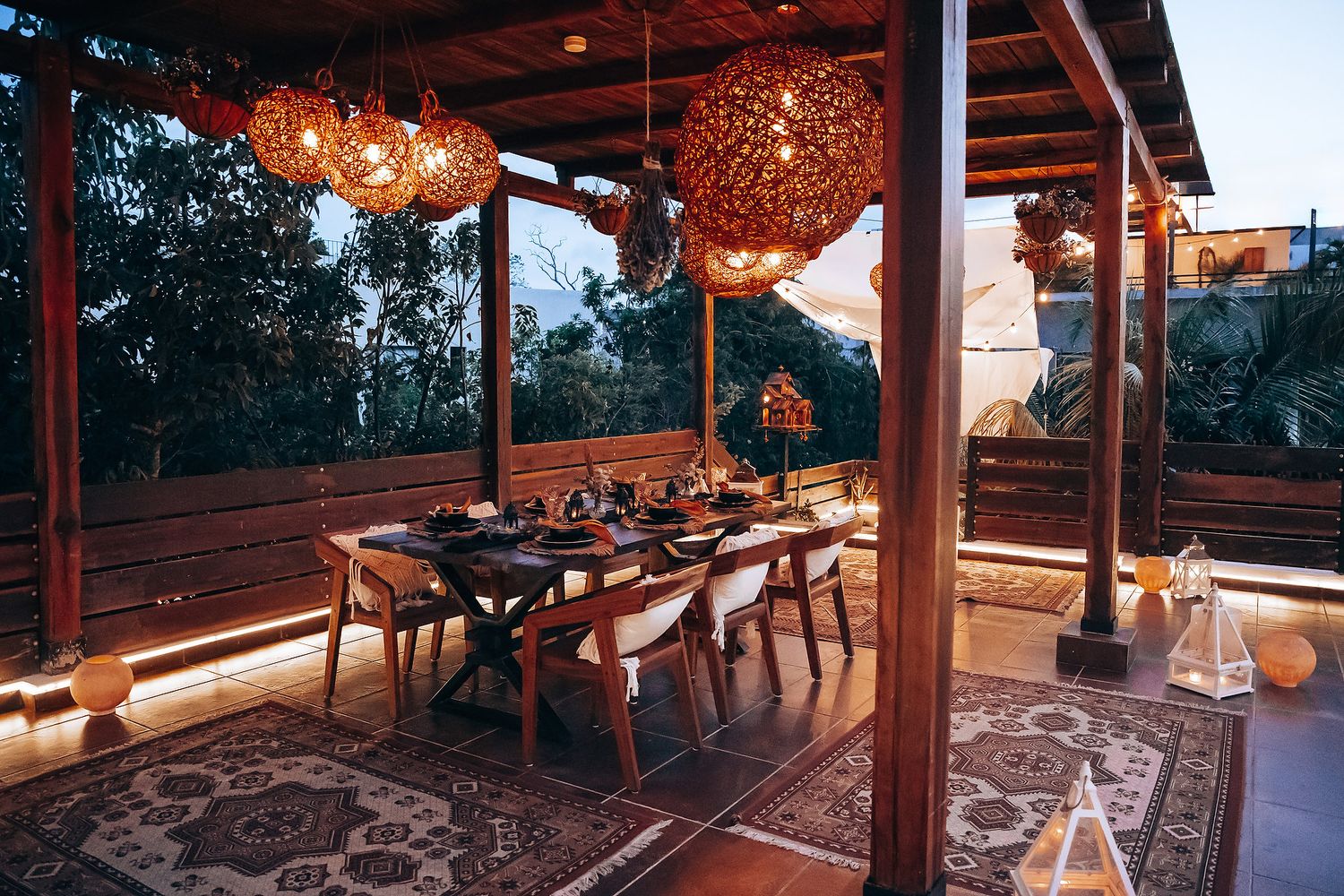 Elegant open-air pavilion dining setup with rattan globe pendant lights over wooden table at twilight.
