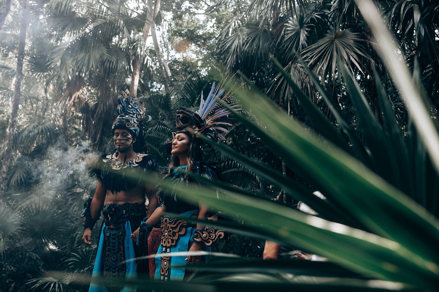 Two people in elaborate Mayan ceremonial costumes with feathered headdresses stand among lush tropical jungle foliage.