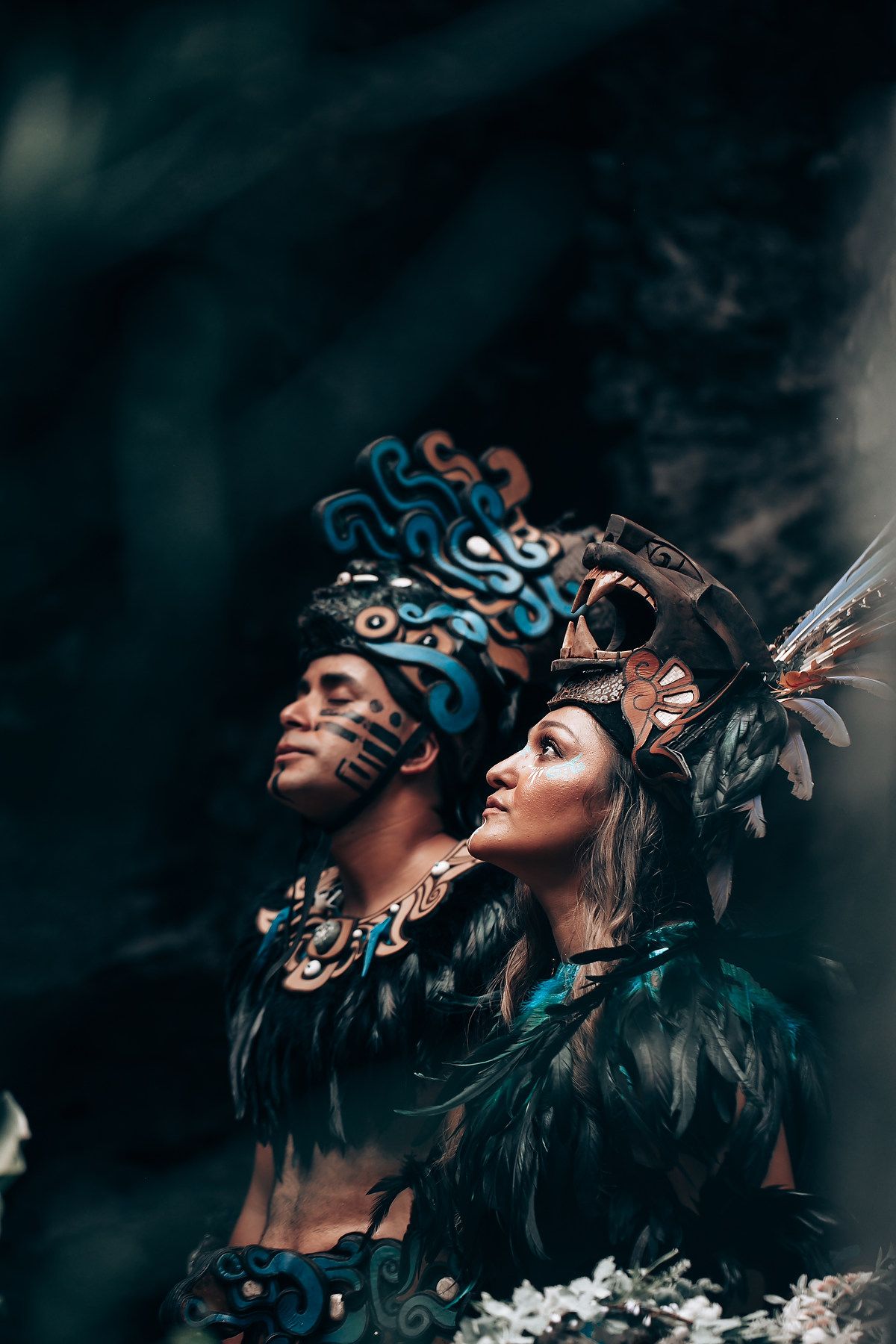 Man and woman in detailed Aztec headdresses and dark feathered costumes gaze upward in a dramatic pose.