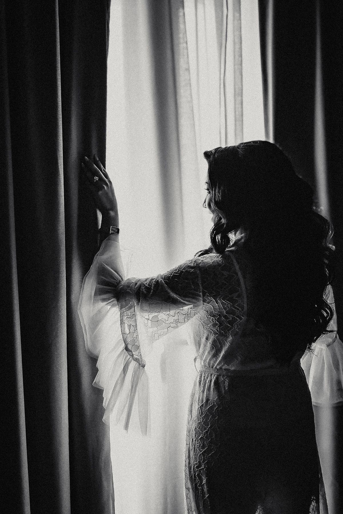 Silhouette of a woman in elegant lace robe reaching toward curtains by a bright window.
