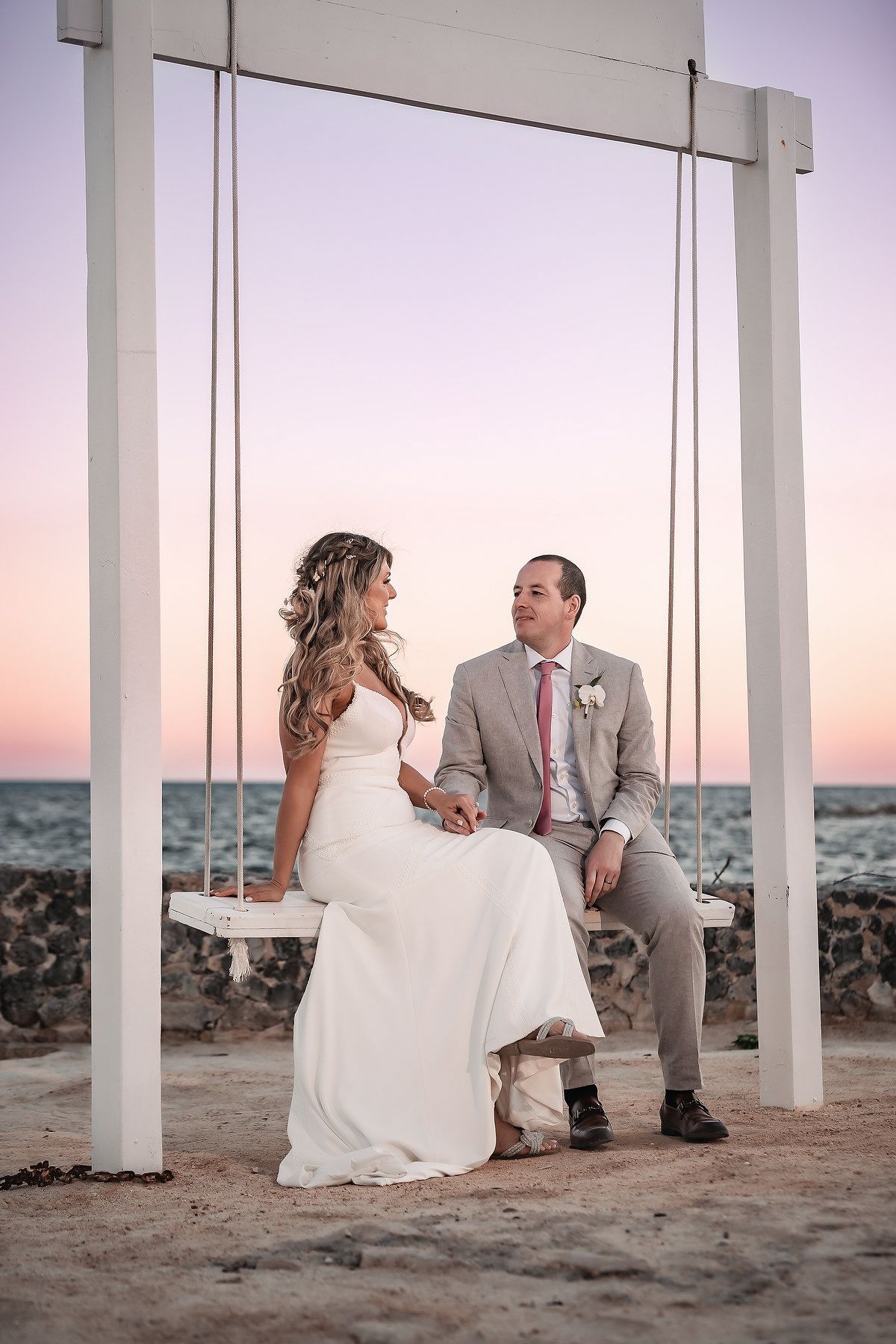 Bride and groom sitting on a white swing by the ocean at sunset, sharing a romantic moment on their wedding day.