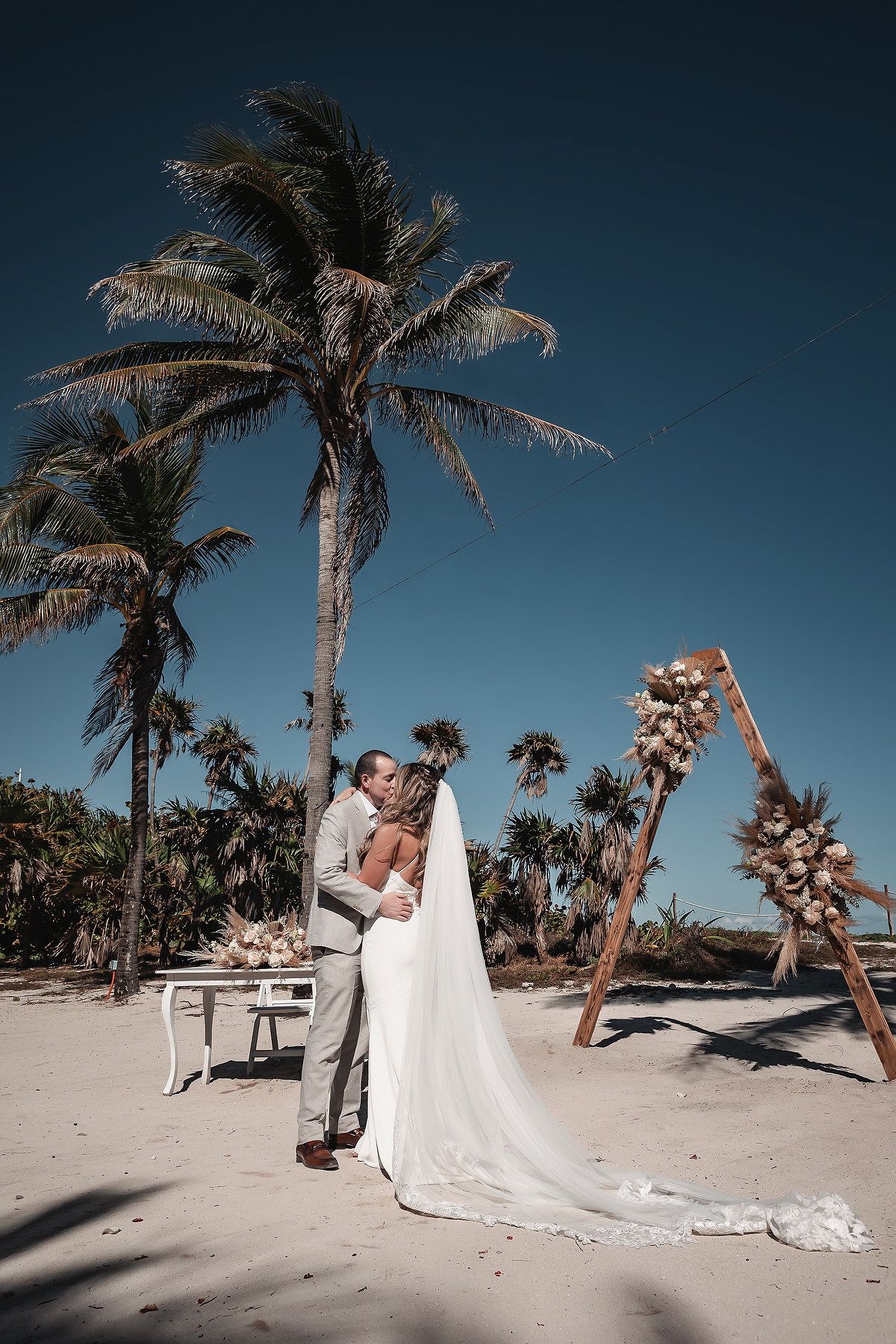 Newlyweds share a romantic kiss on a sandy beach surrounded by palm trees after their outdoor wedding.