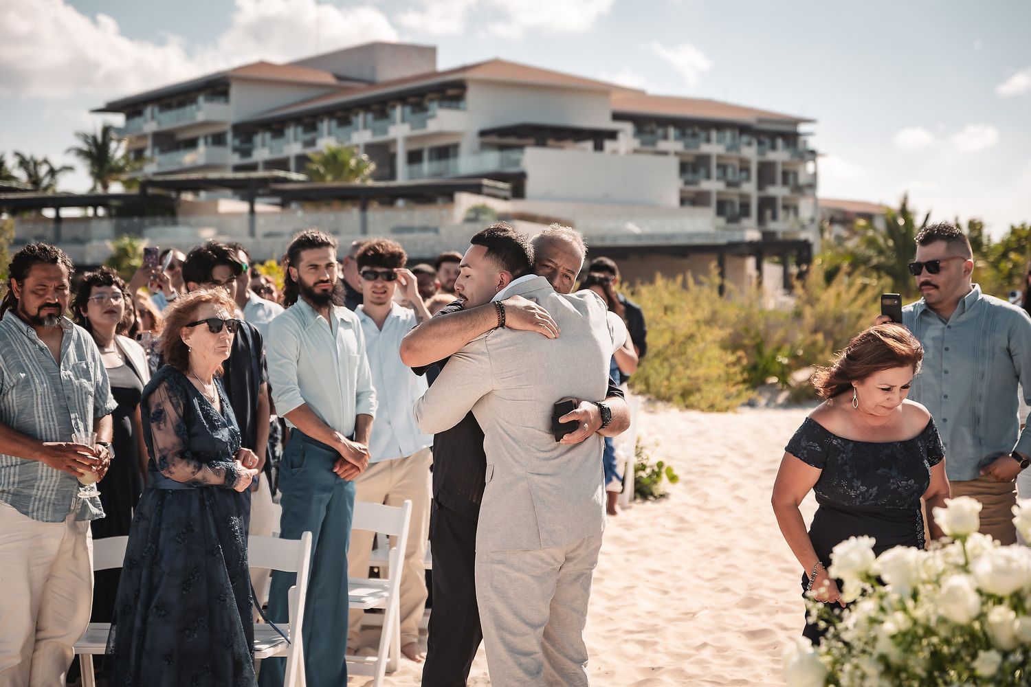 Two men share an emotional hug at a beachside wedding ceremony with guests watching and a resort in the background.