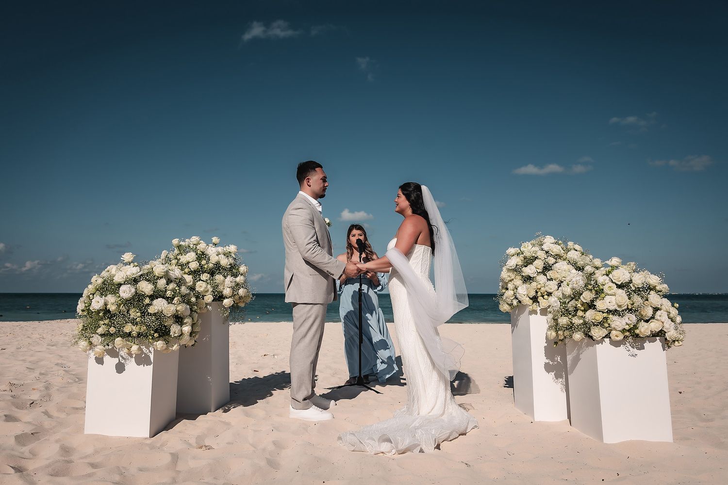 Couple exchanging vows on a beach wedding ceremony with white floral arrangements and an officiant under a blue sky.