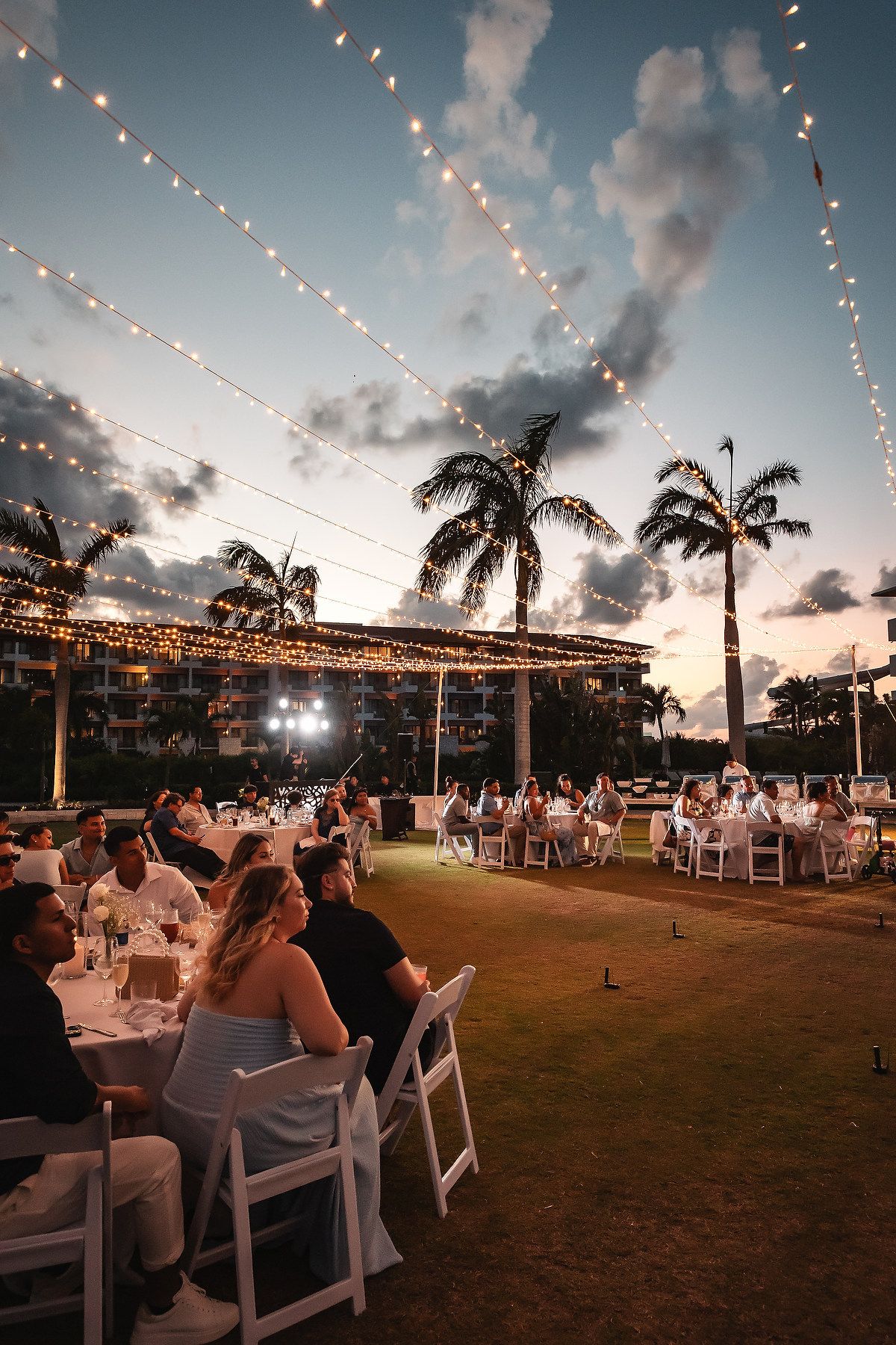 Outdoor wedding reception at sunset with string lights, white chairs, and palm trees under a colorful sky.