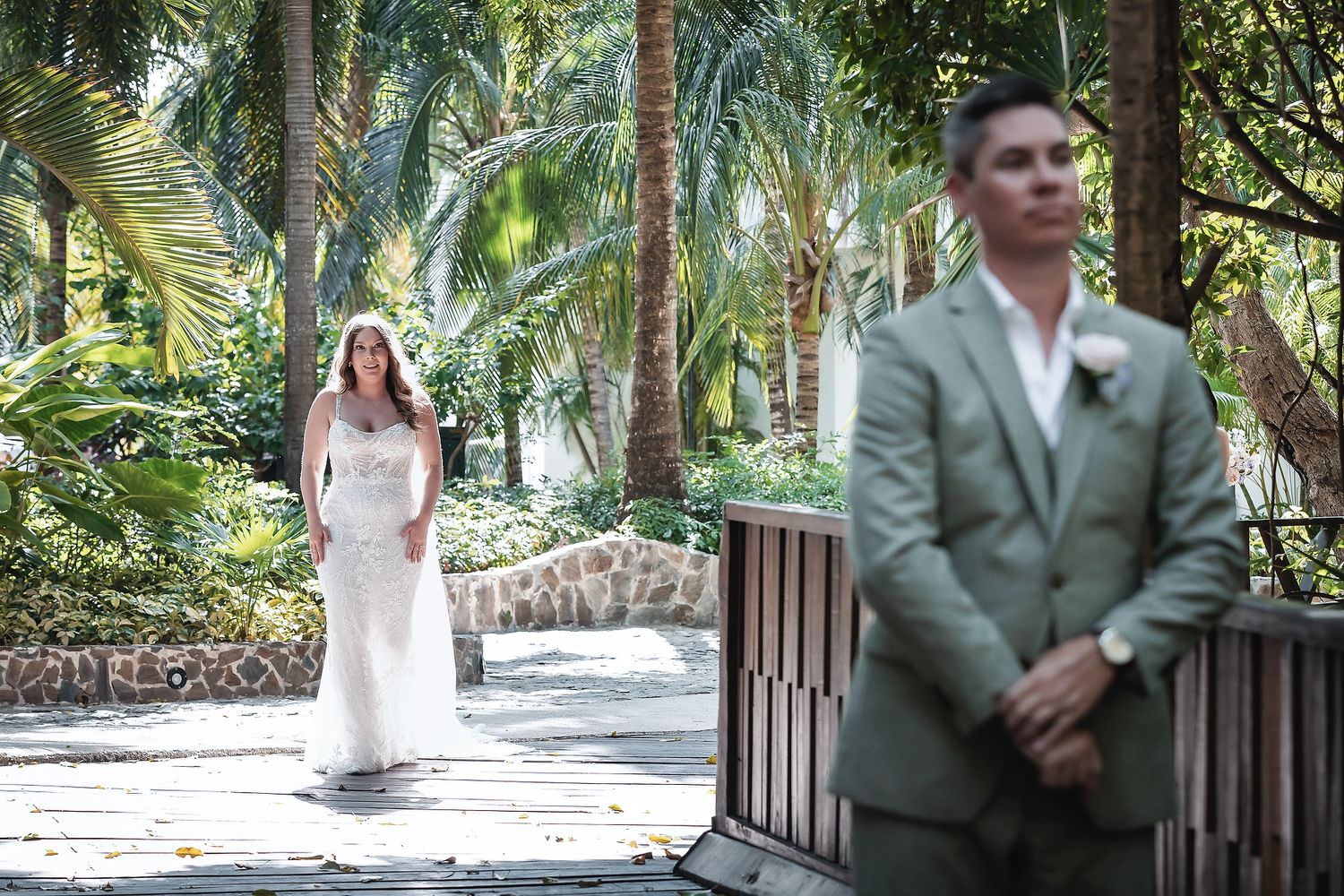 Bride in white gown walks toward groom in sage suit during outdoor tropical garden wedding ceremony.