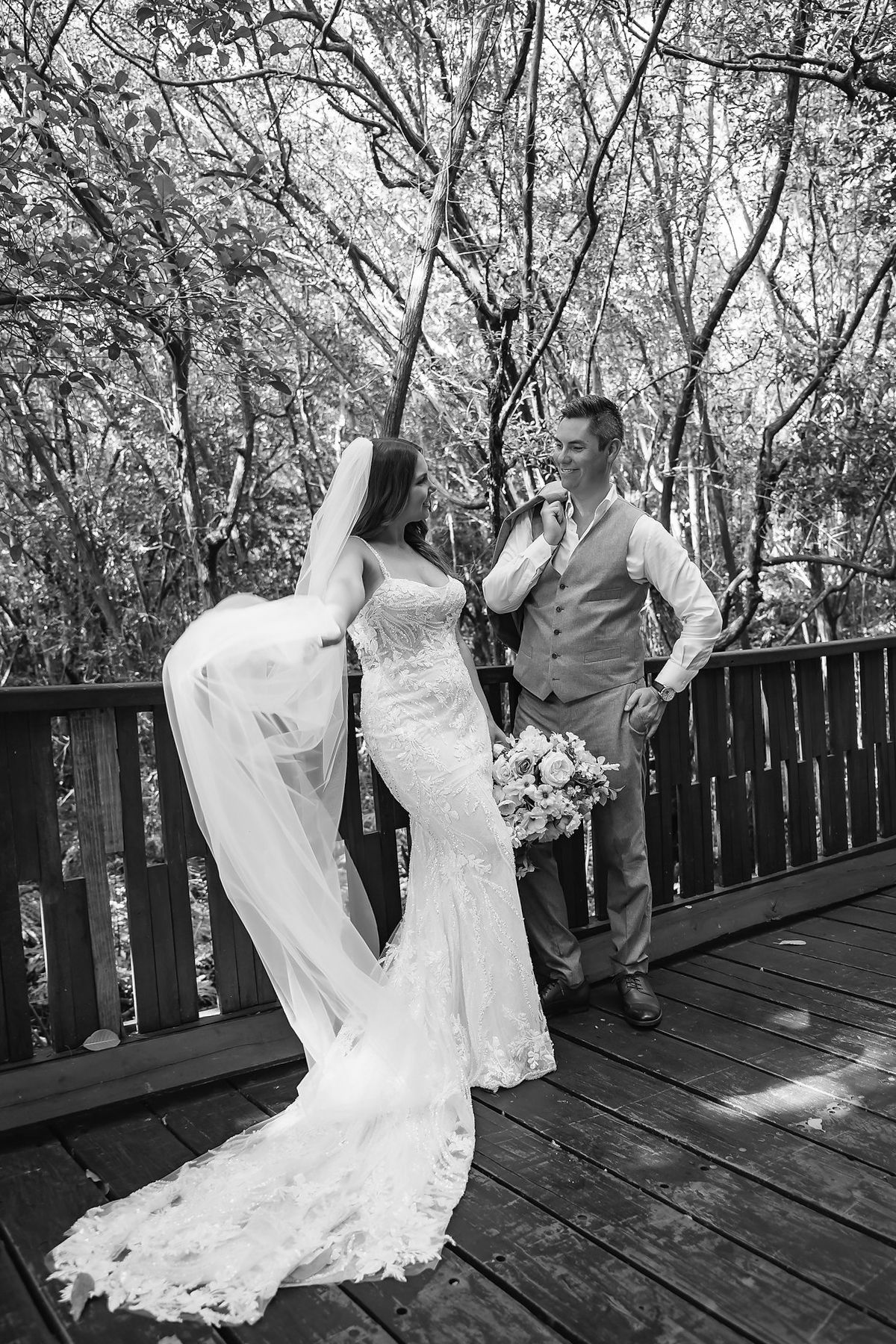 Black and white photo of a bride and groom sharing a romantic moment on a wooden deck surrounded by trees.
