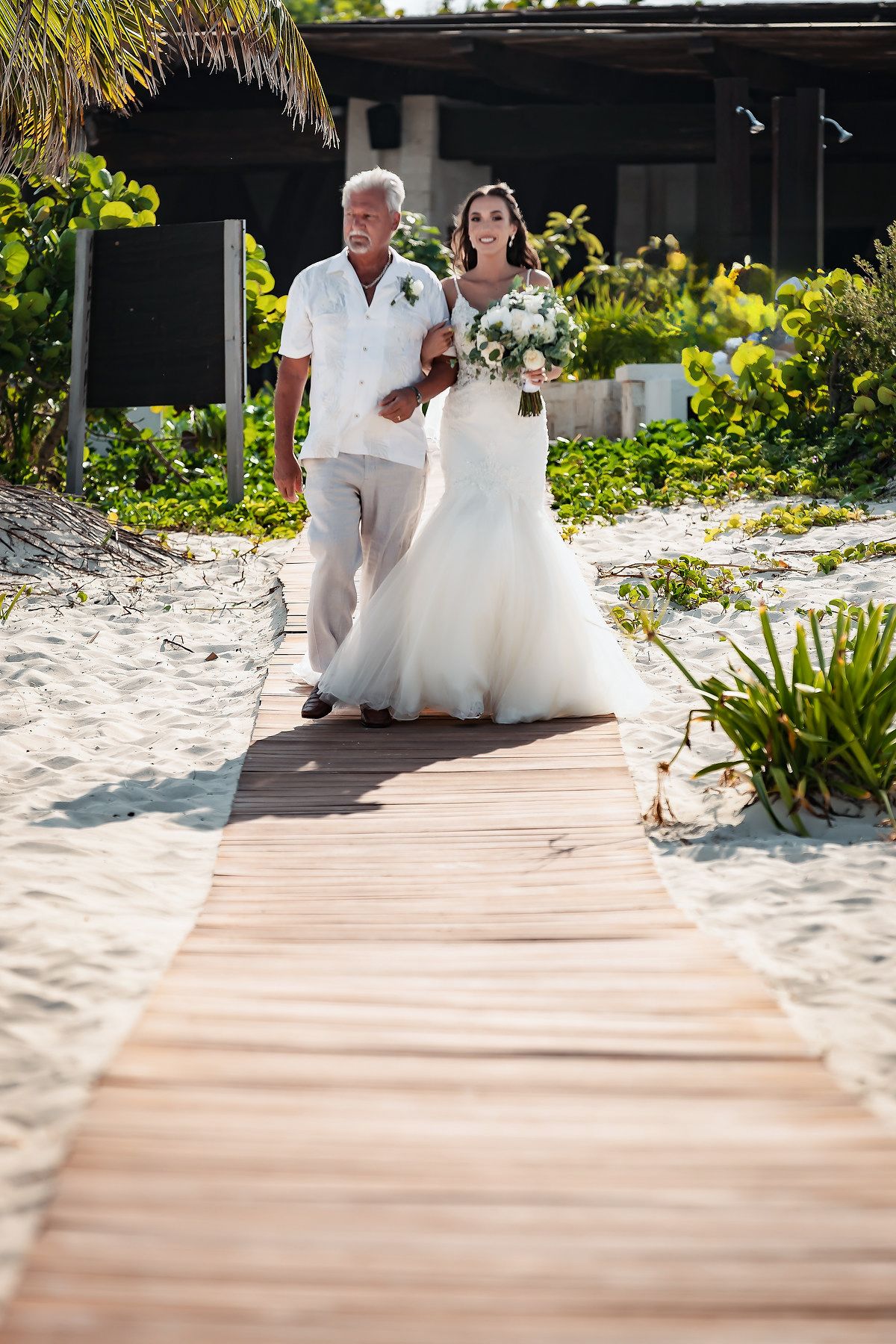 Bride in white mermaid gown walks with older man on wooden beach boardwalk, holding white bouquet at tropical wedding.