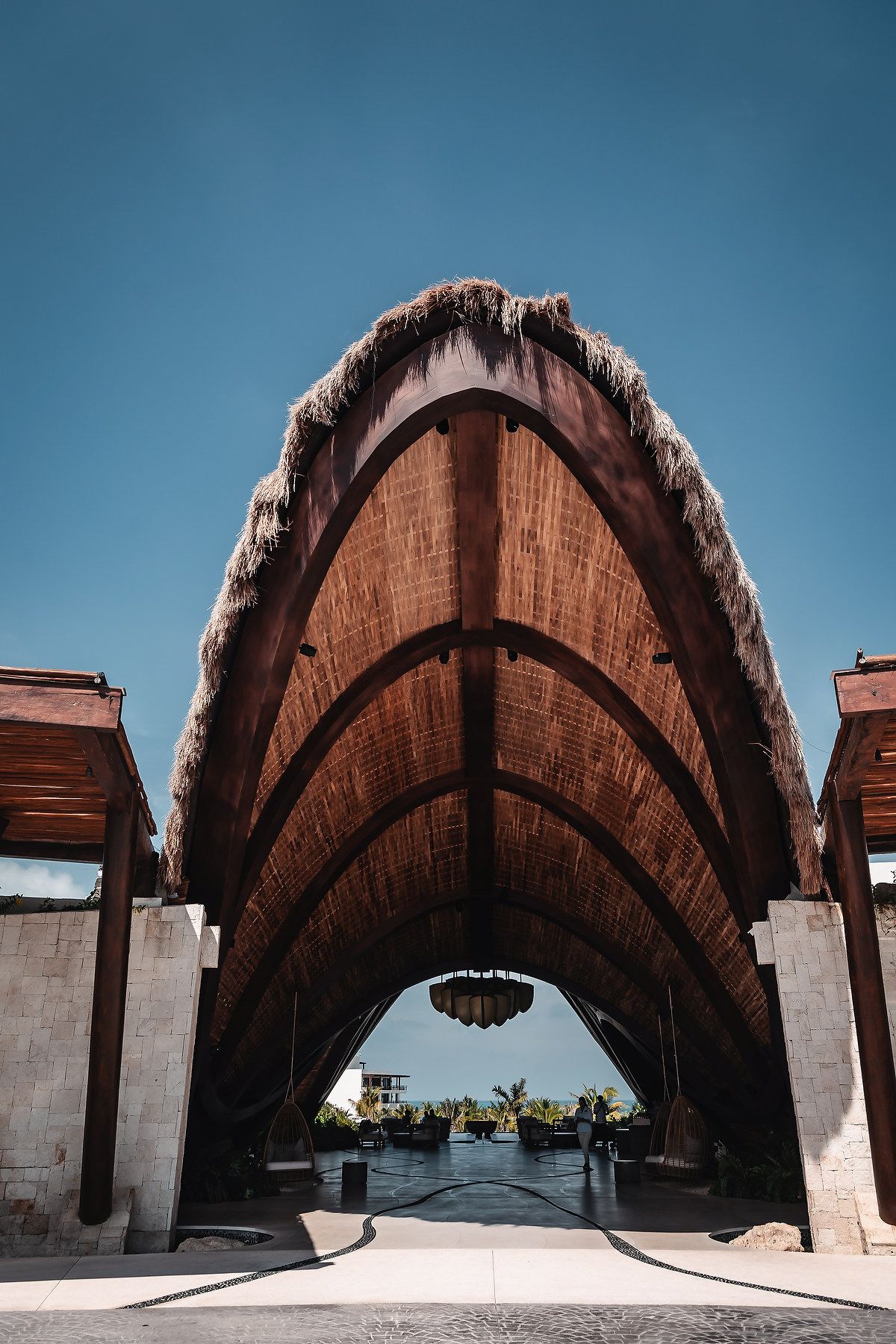 Stunning arched wooden palapa entrance with thatched roof framing a tropical resort view under clear blue sky.
