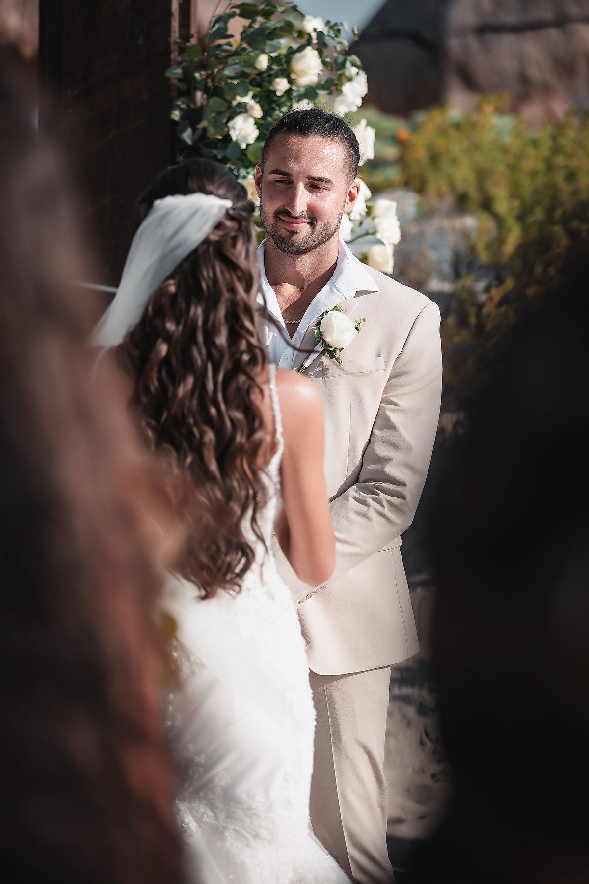 Groom in beige suit smiling at bride during outdoor wedding ceremony with white floral arch backdrop.