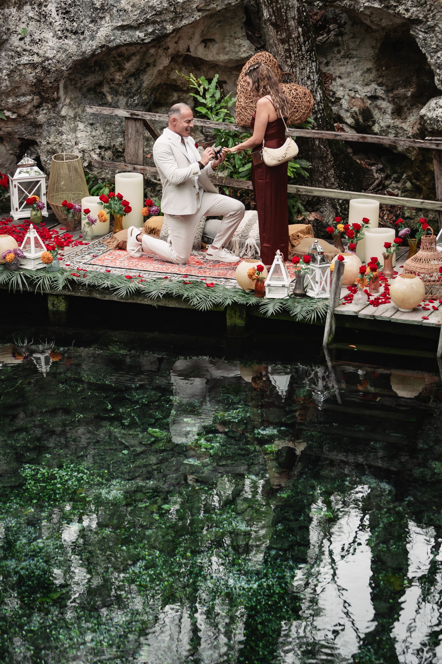 Man proposes on decorated dock over serene water, surrounded by candles, red roses, and tropical decor in a cave setting.