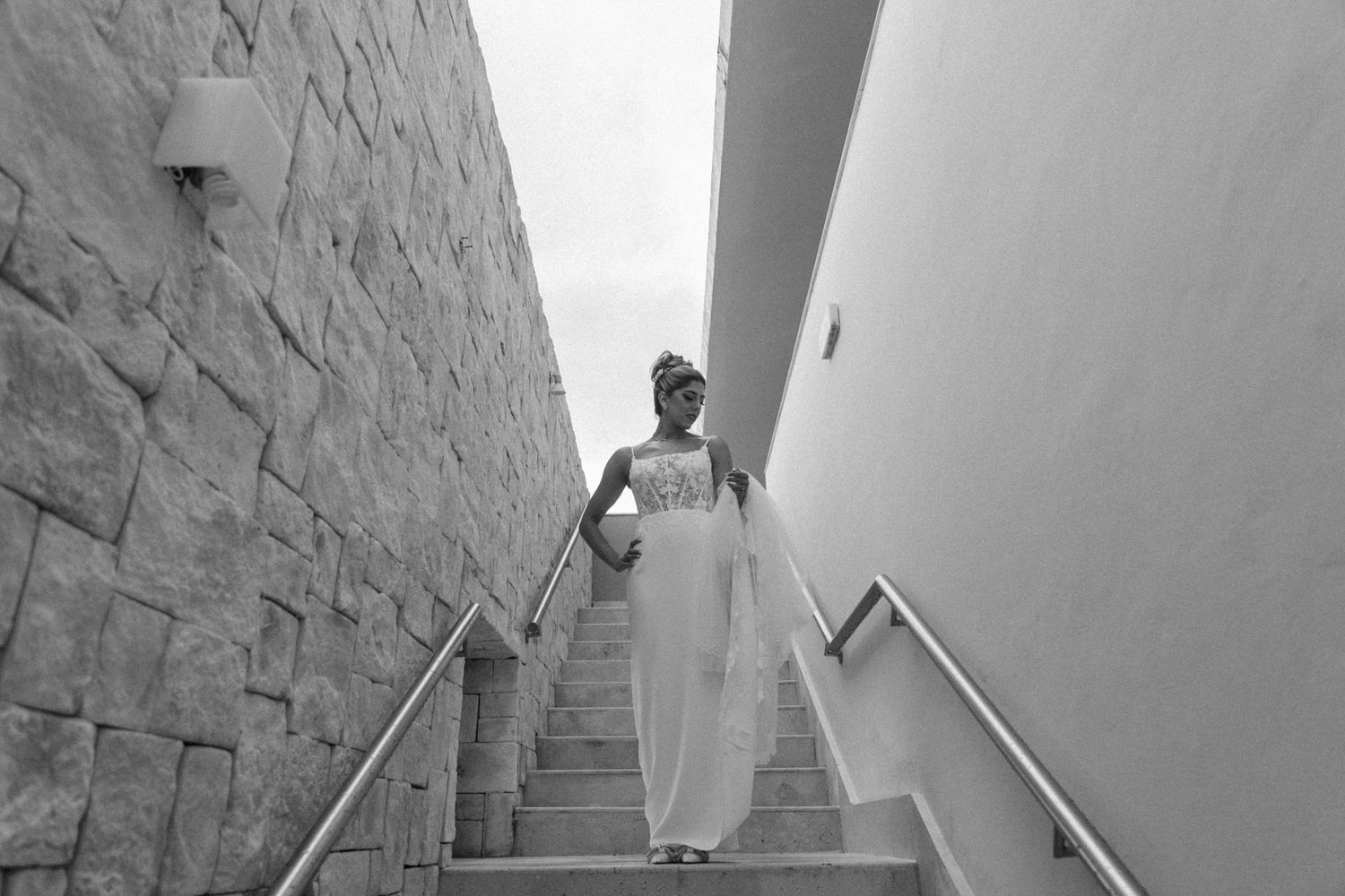 Bride in white gown descending outdoor stone staircase with metal railings in black and white photo.