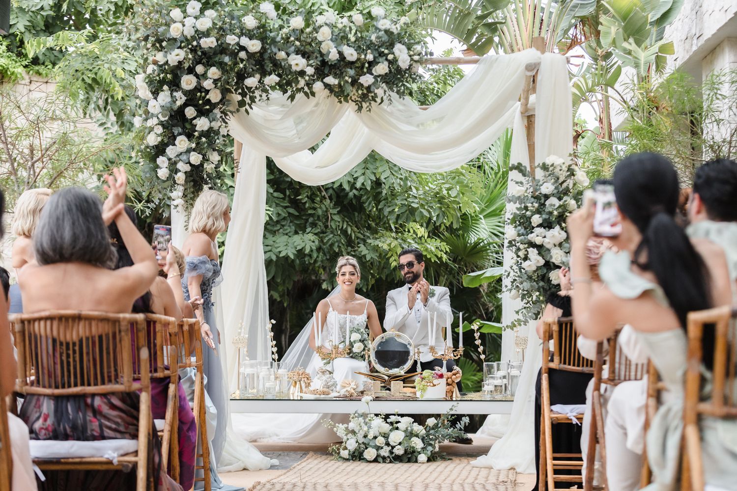 Couple exchanges vows under floral arch with white draping at elegant outdoor wedding ceremony surrounded by lush greenery.