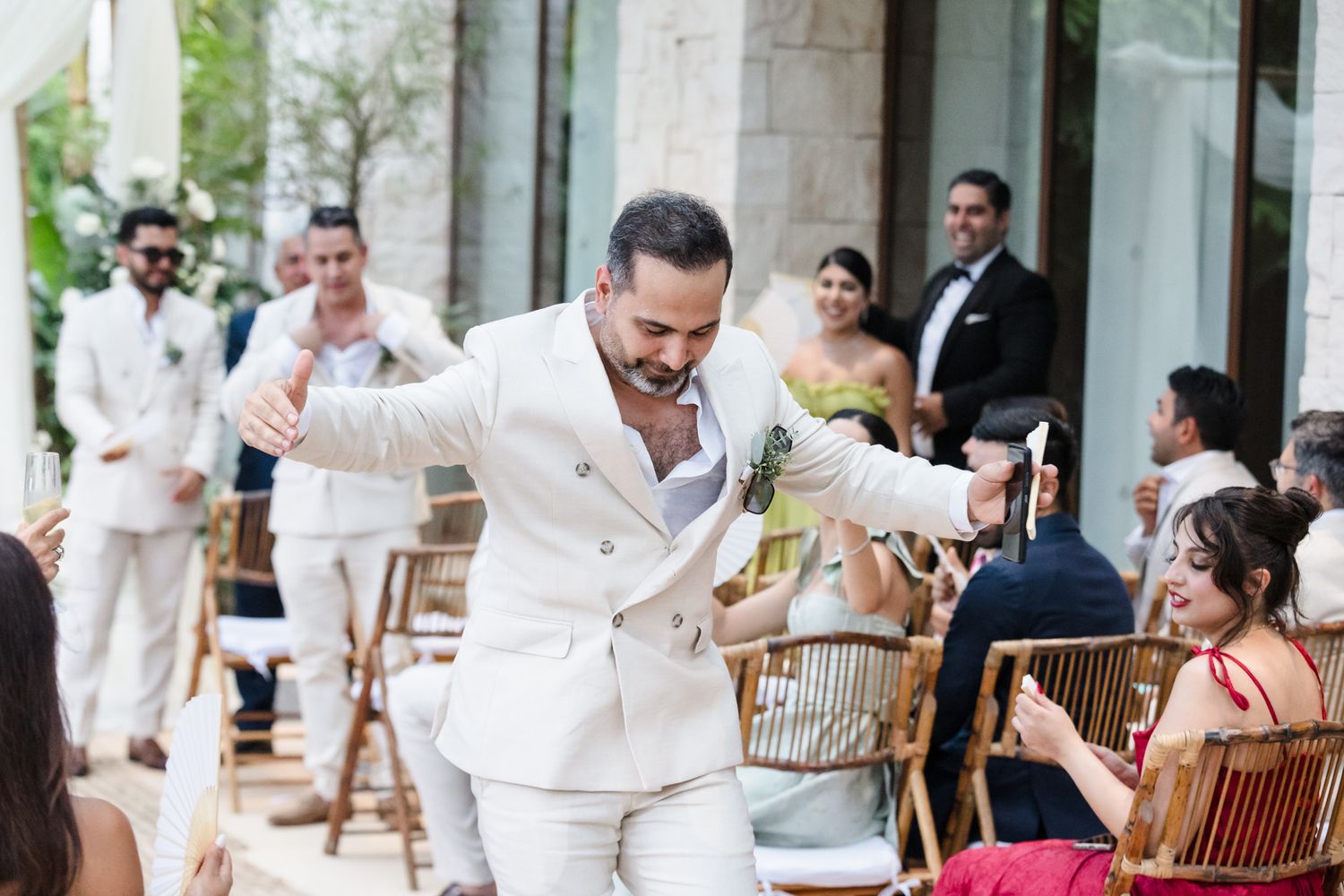 Groom in white suit dancing joyfully at outdoor wedding ceremony with guests seated and celebrating.