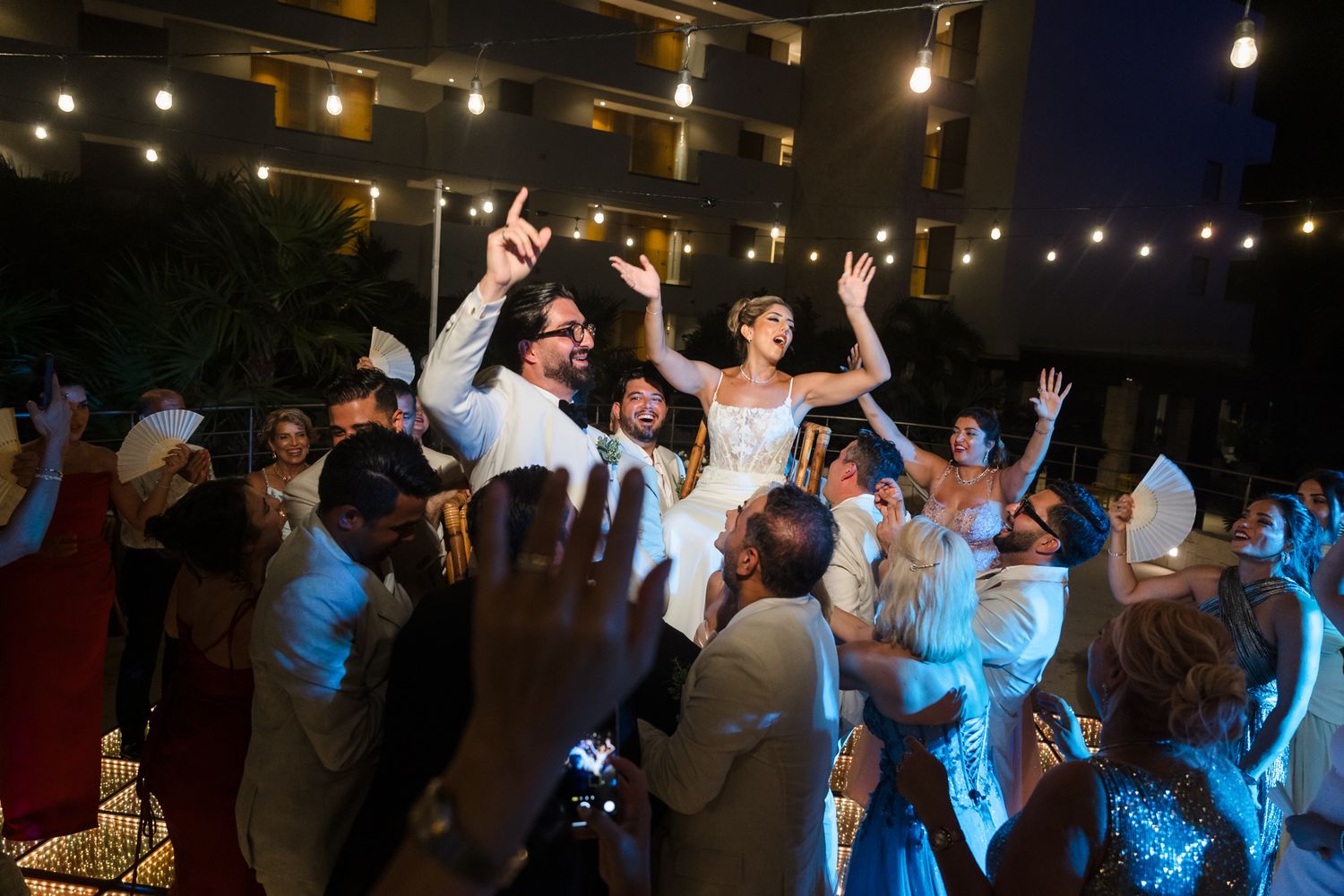 Bride and groom lifted by guests at outdoor wedding reception with string lights at night.