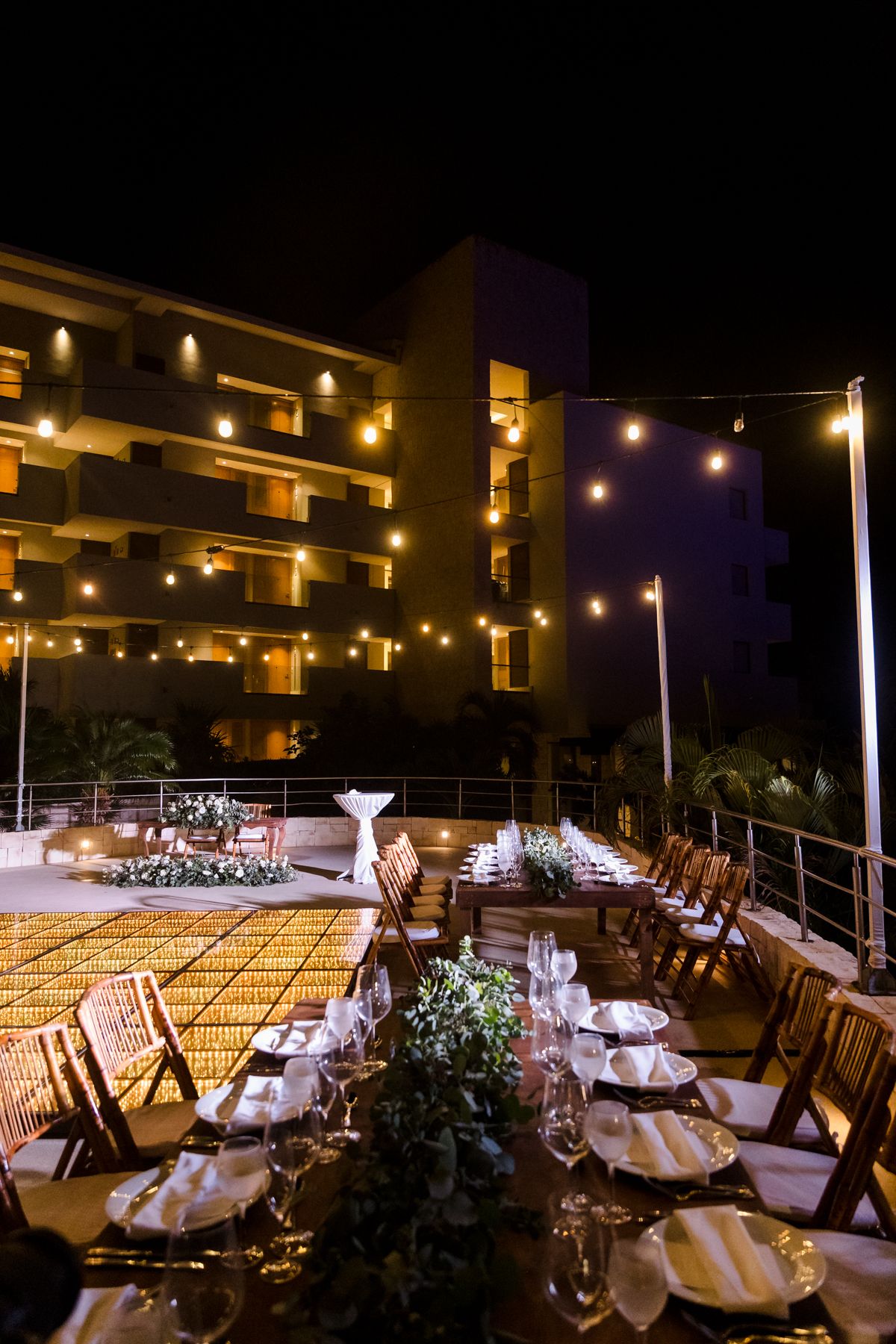 Elegant outdoor rooftop wedding reception set at night with string lights, long dining tables, and glowing hotel backdrop.