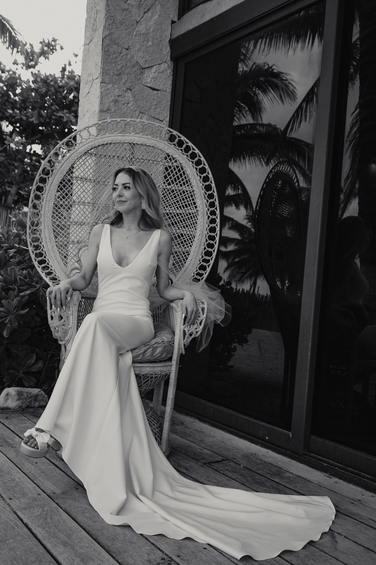 Elegant bride in white gown seated in wicker peacock chair on wooden deck with tropical backdrop, black and white photo.