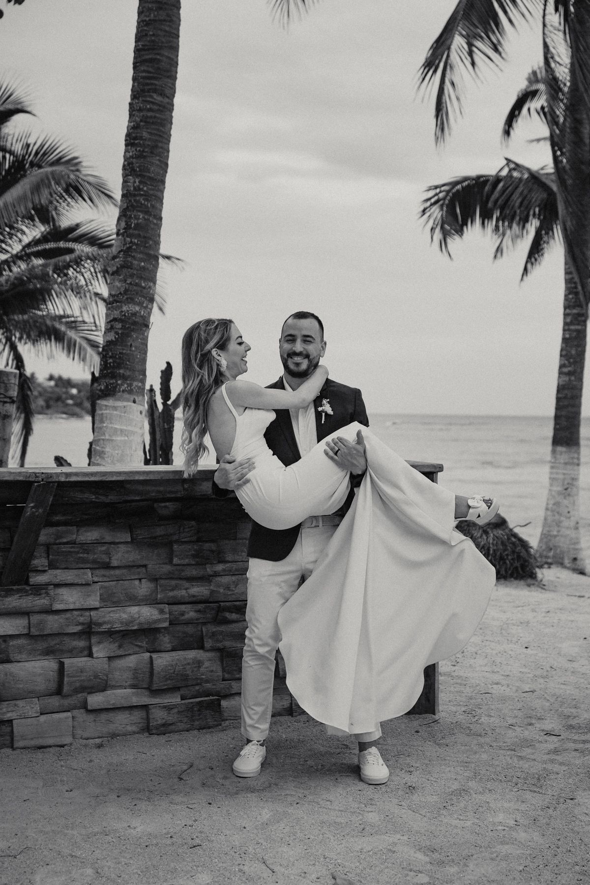 Black and white photo of groom lifting smiling bride in wedding dress on a sandy beach with palm trees and ocean.