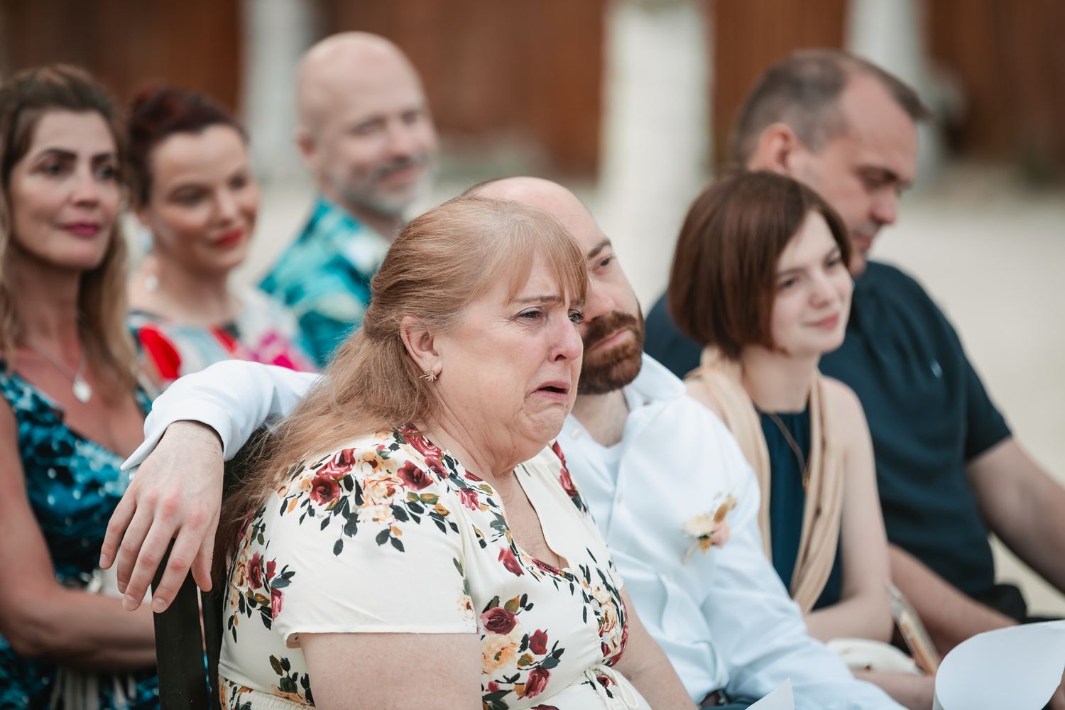 Emotional wedding guest with teary eyes seated among other attendees during an outdoor beach ceremony.