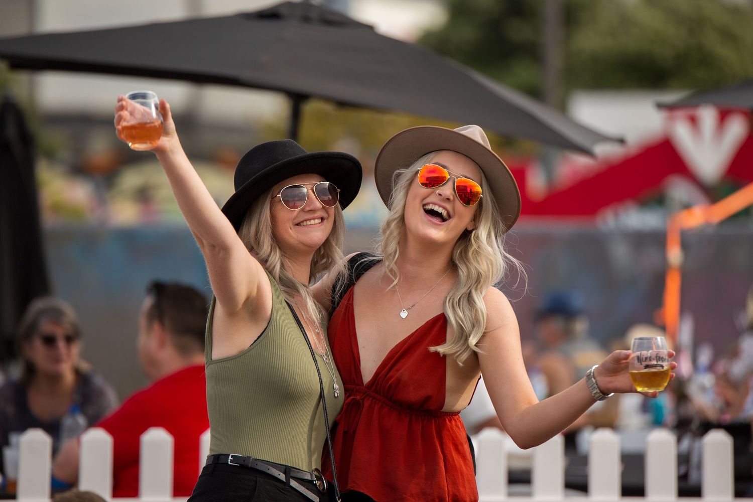 Two friends in summer outfits enjoying drinks and laughing at an outdoor festival.