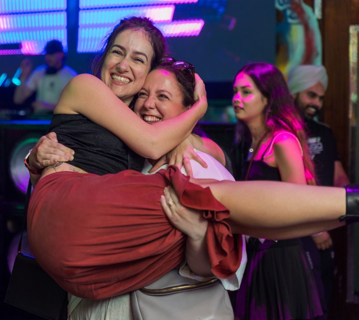 Two friends sharing a joyful embrace on a dance floor with colorful lighting in the background.