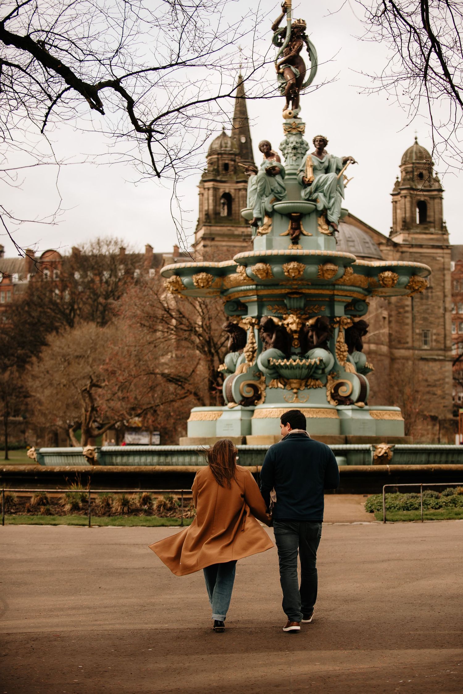 Two people walk hand in hand near an ornate fountain in a park on an autumn day with bare trees in the background.