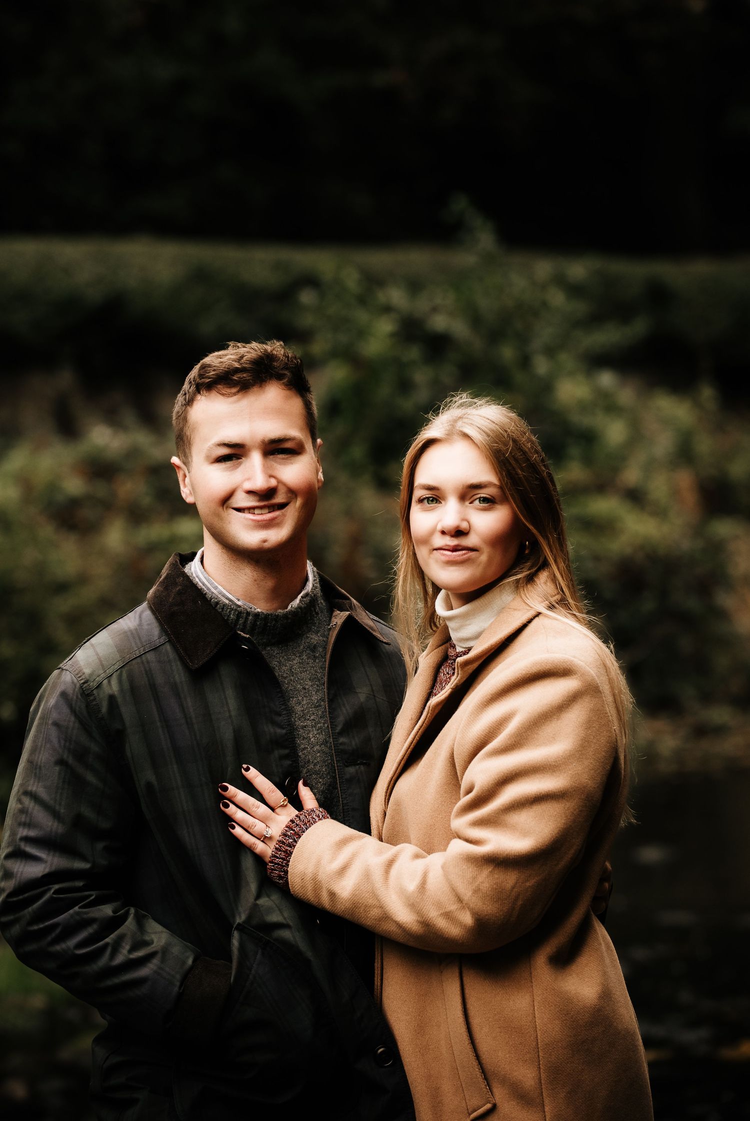 Couple in casual winter coats posing together outdoors near a lake.