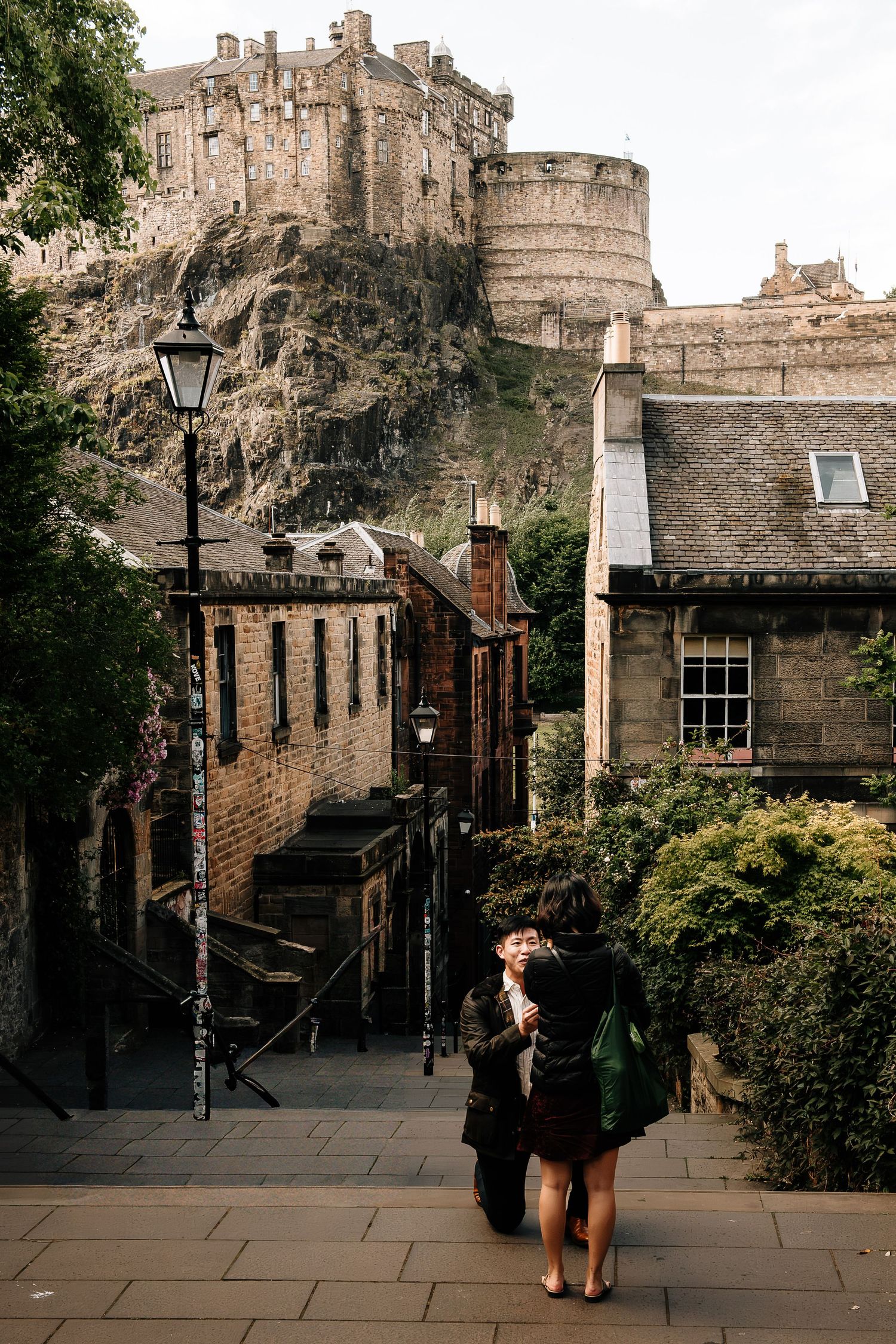 Couple embracing on cobblestone steps below Edinburgh Castle, historic stone buildings and lush greenery surrounding them.
