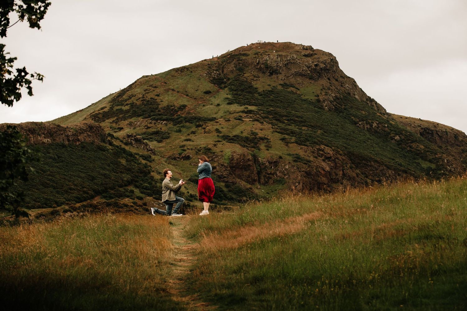 Two hikers on a grassy trail with Arthur's Seat rocky hill in the background under overcast skies.