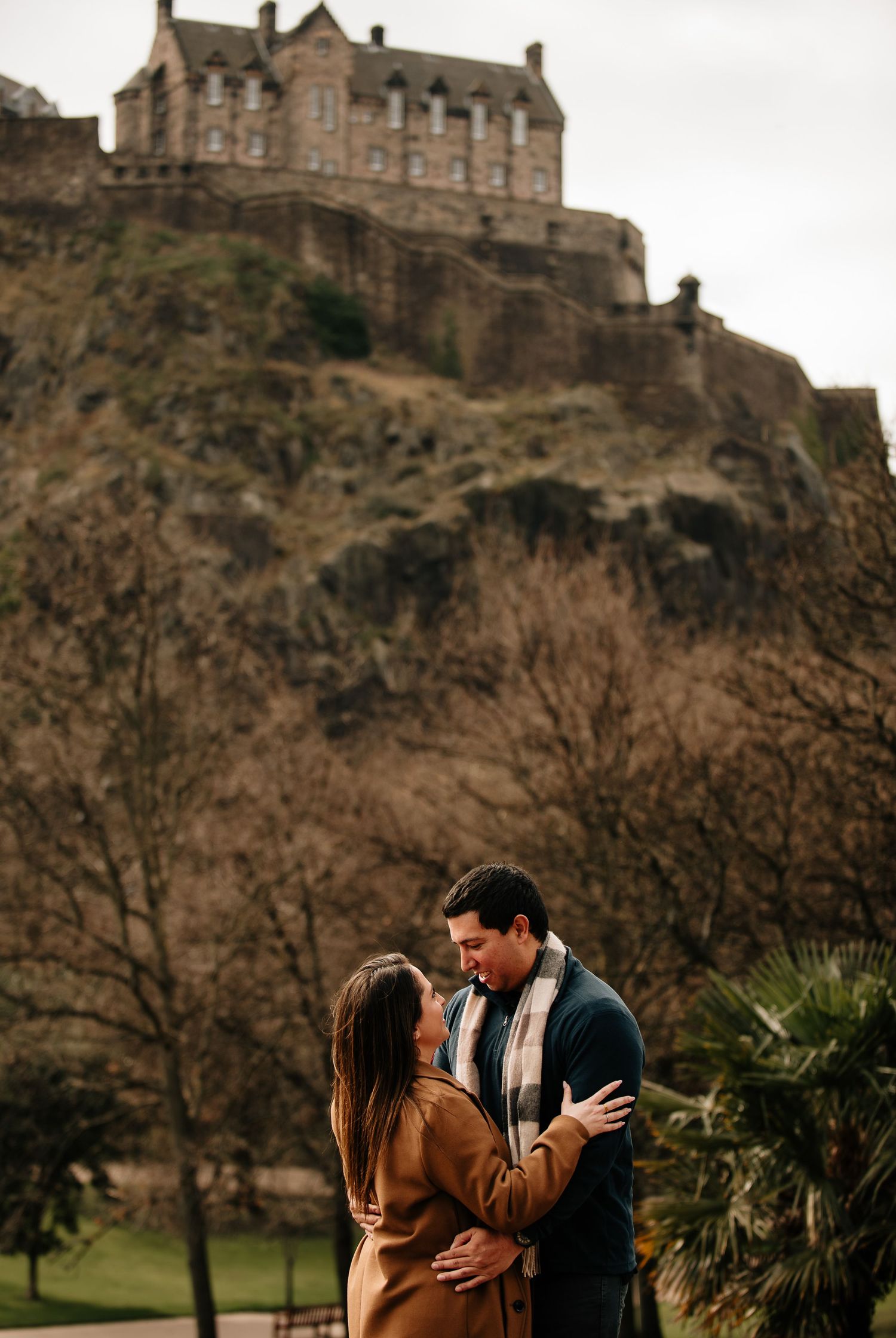 Edinburgh Castle stands majestically on a rocky hill while a couple shares a romantic moment in Princes Street Gardens below.