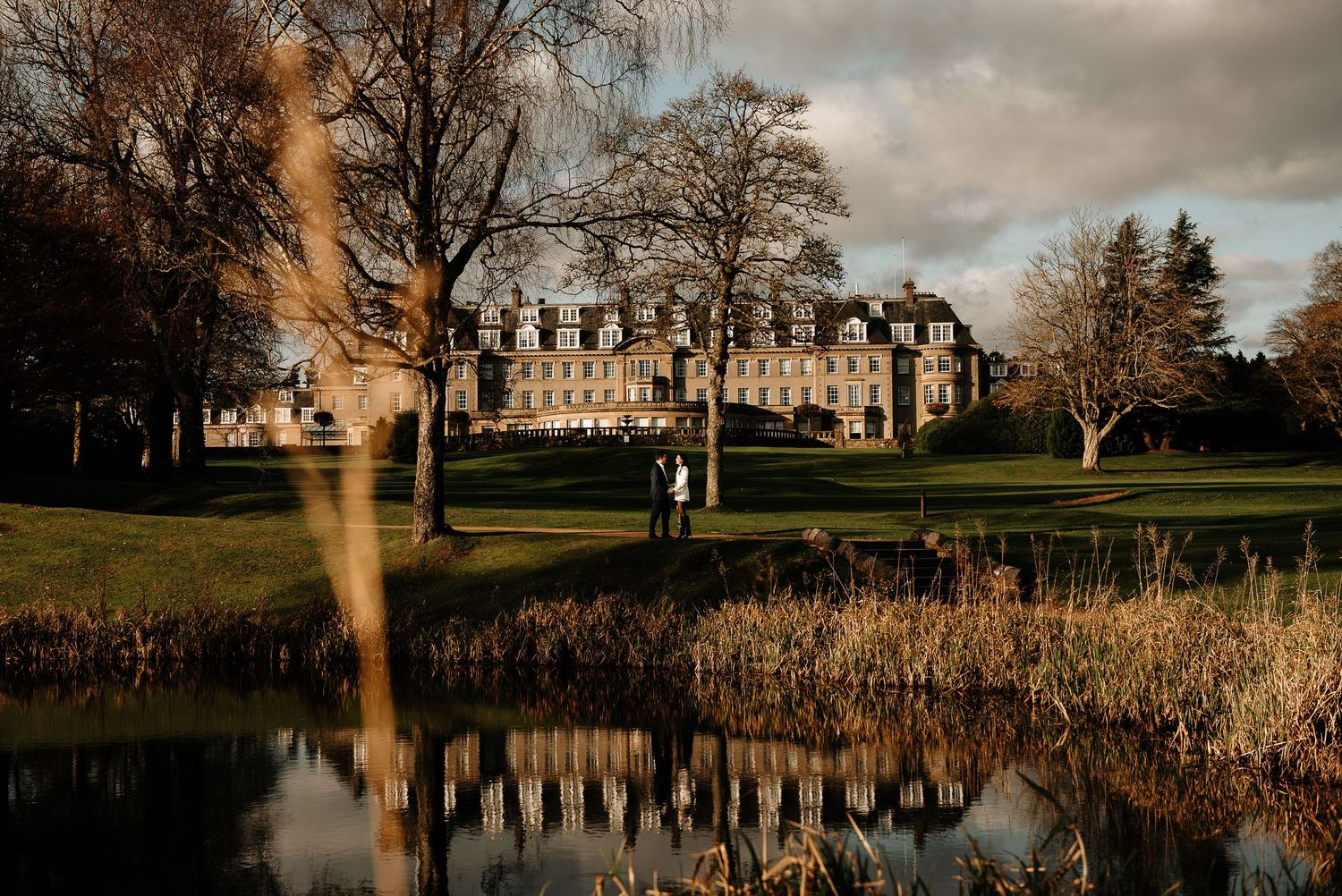 A couple stands silhouetted against a grand manor house reflected in a pond at sunset.