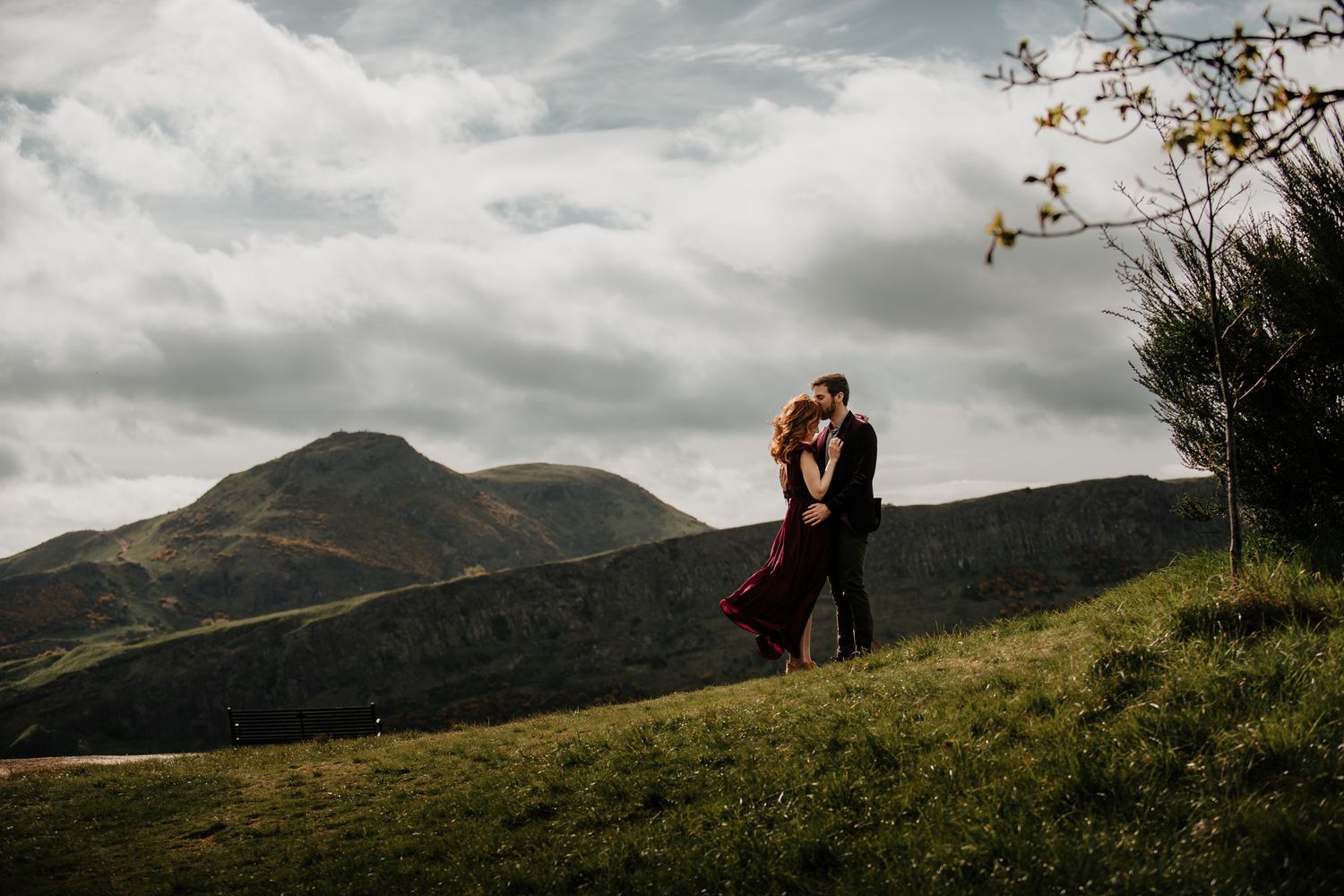 A couple embraces on a grassy hilltop against a dramatic Arthur's Seat, with moody cloudy skies.