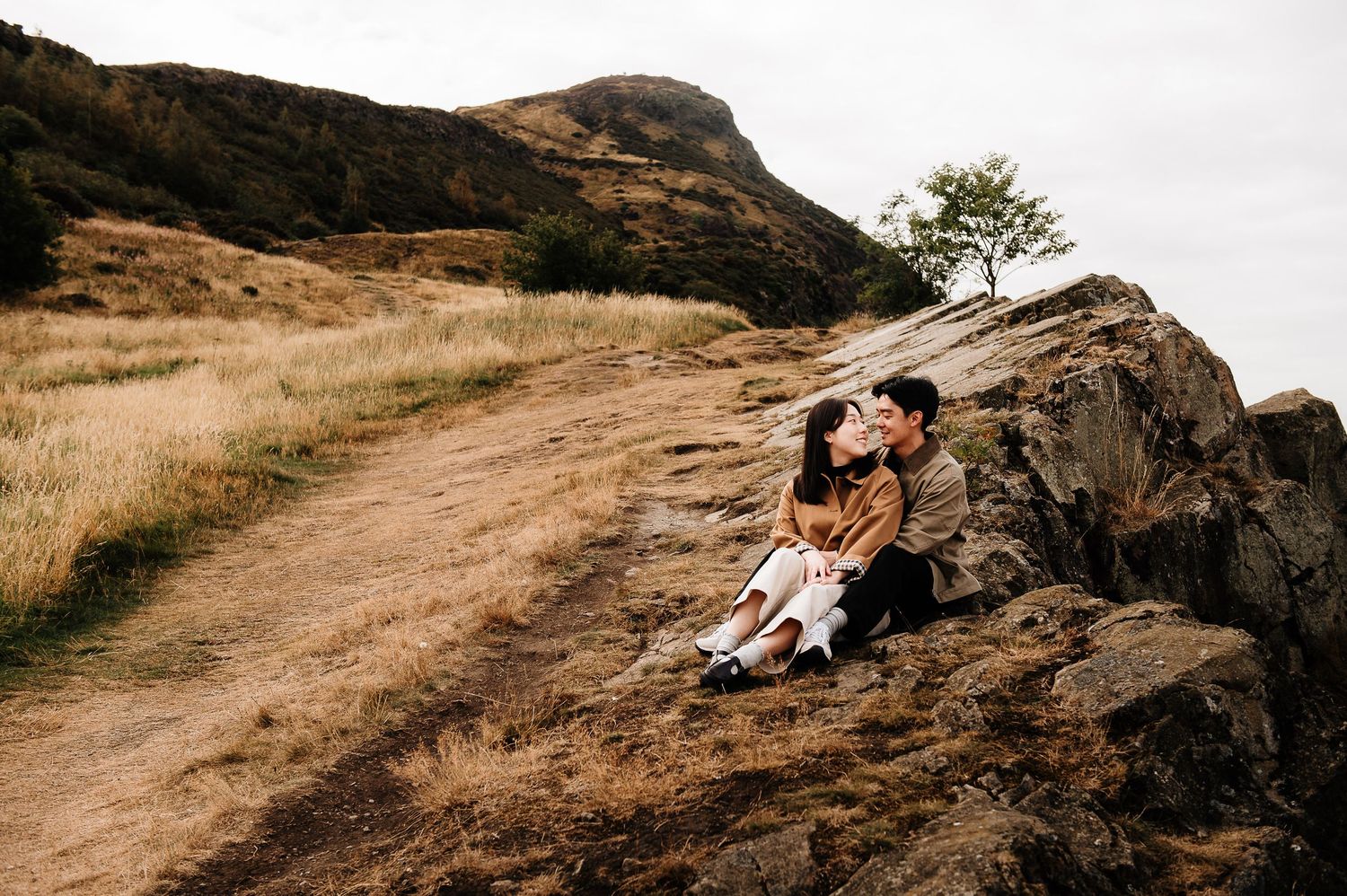 A couple shares a romantic moment while sitting on a rocky cliff overlooking Holyrood Park.