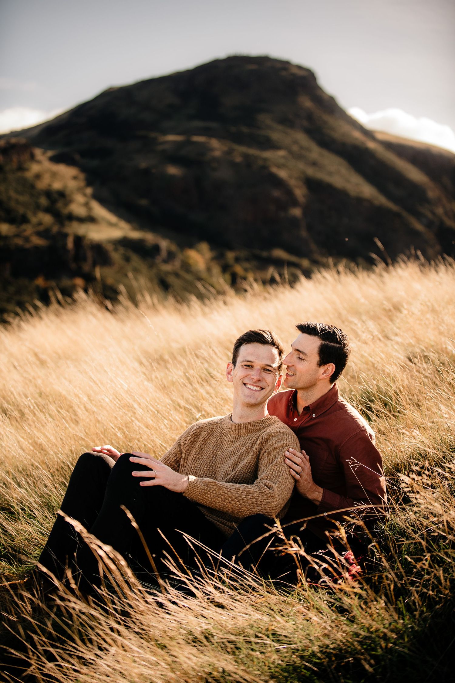 A couple sits in tall golden grass with a mountain silhouetted against the sky.