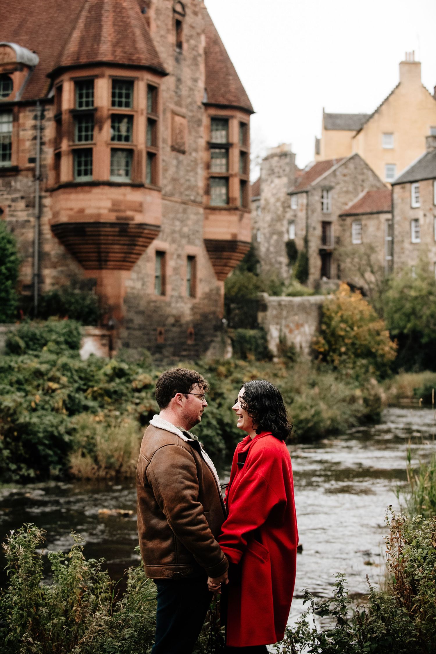 A couple shares a romantic moment by a river with historic stone buildings and architecture behind them.