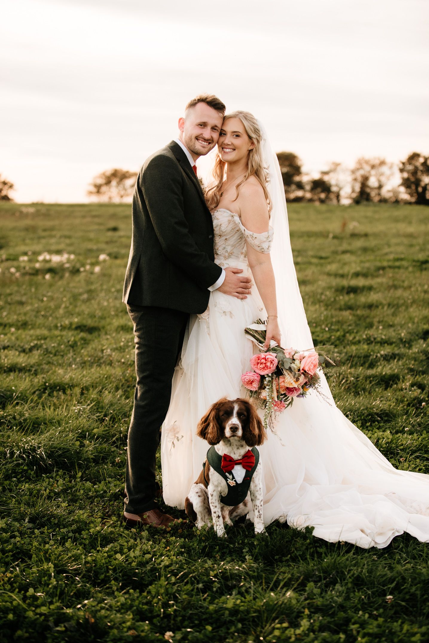 Bride and groom smiling outdoors at sunset with their Springer Spaniel dog wearing a floral collar on their wedding day.