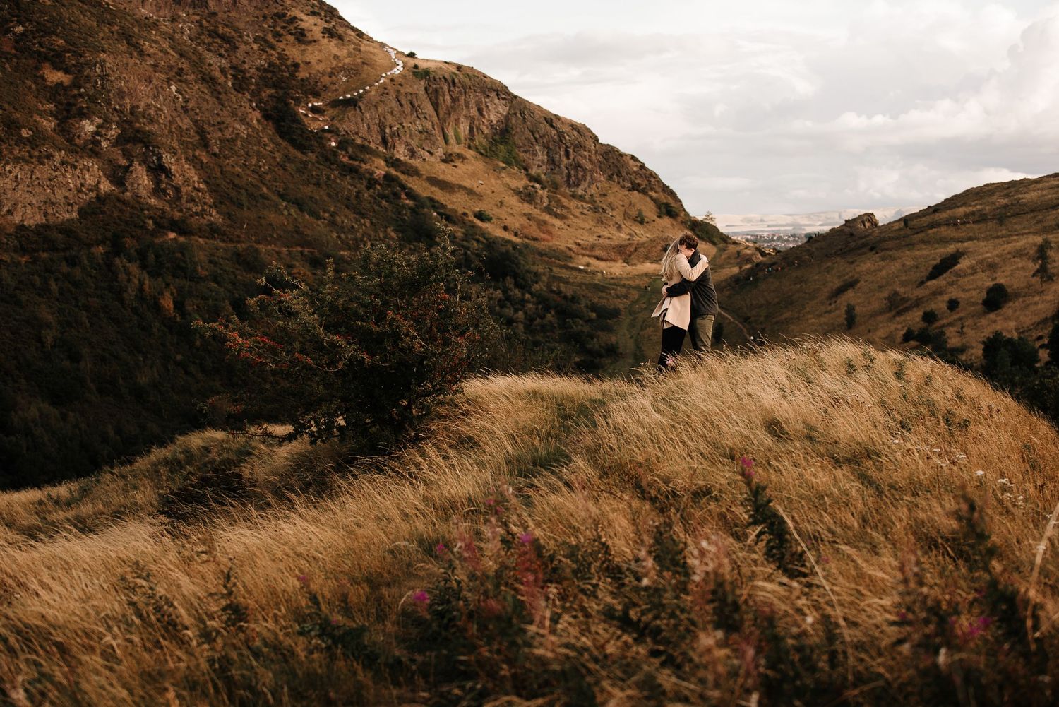 Panoramic view of hikers walking along a winding mountain trail through golden grass.