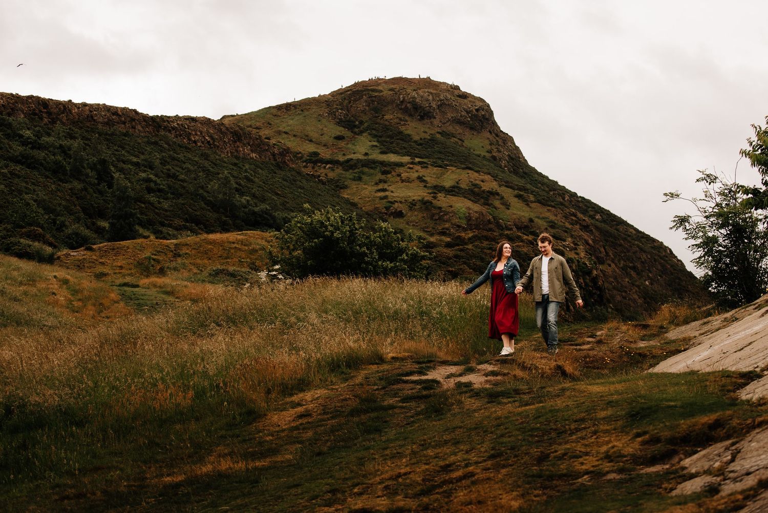 Couple walking hand in hand on a grassy hillside path with a dramatic rocky hill in the background.