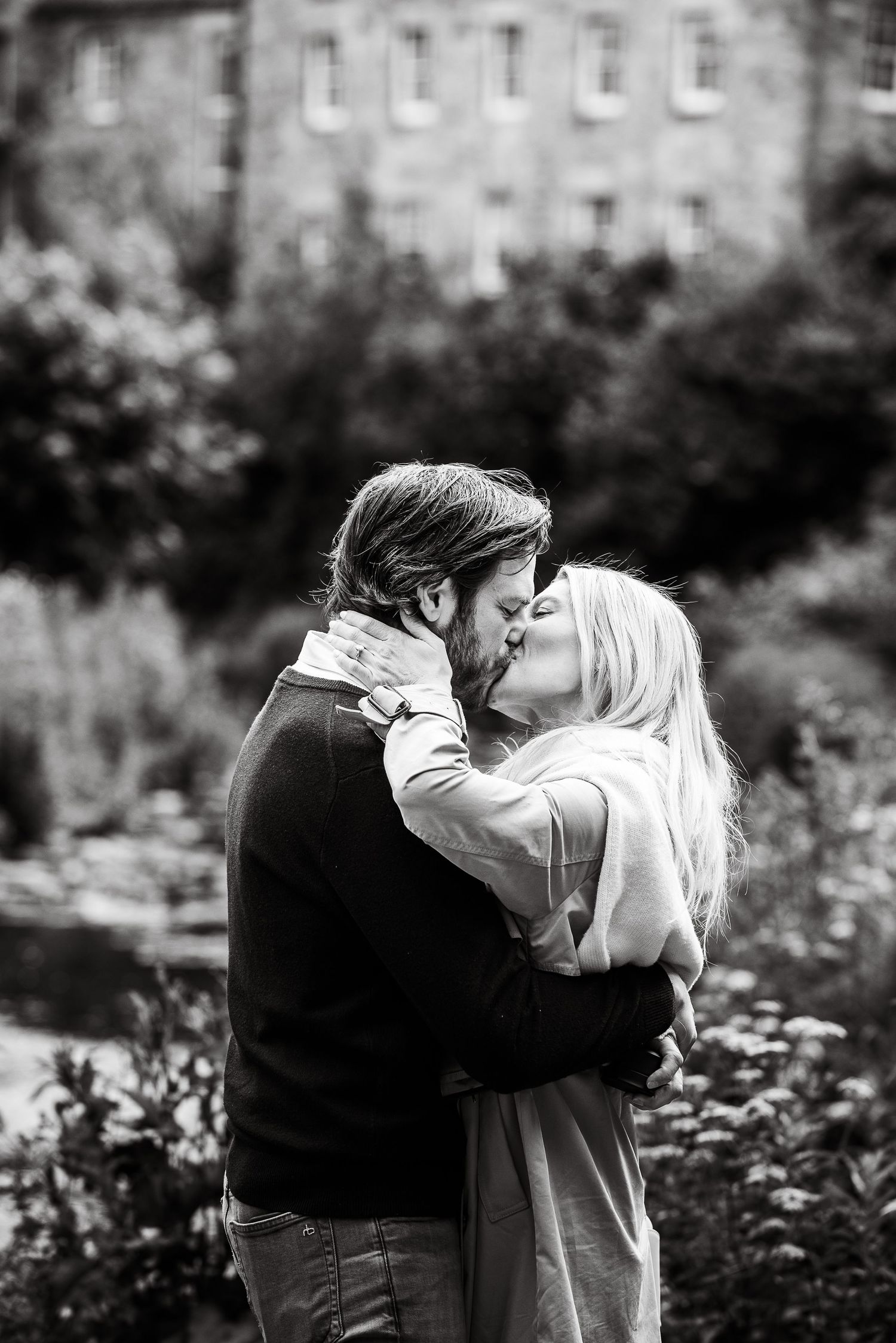 Black and white photo of a romantic couple kissing and embracing outdoors in a lush garden setting.