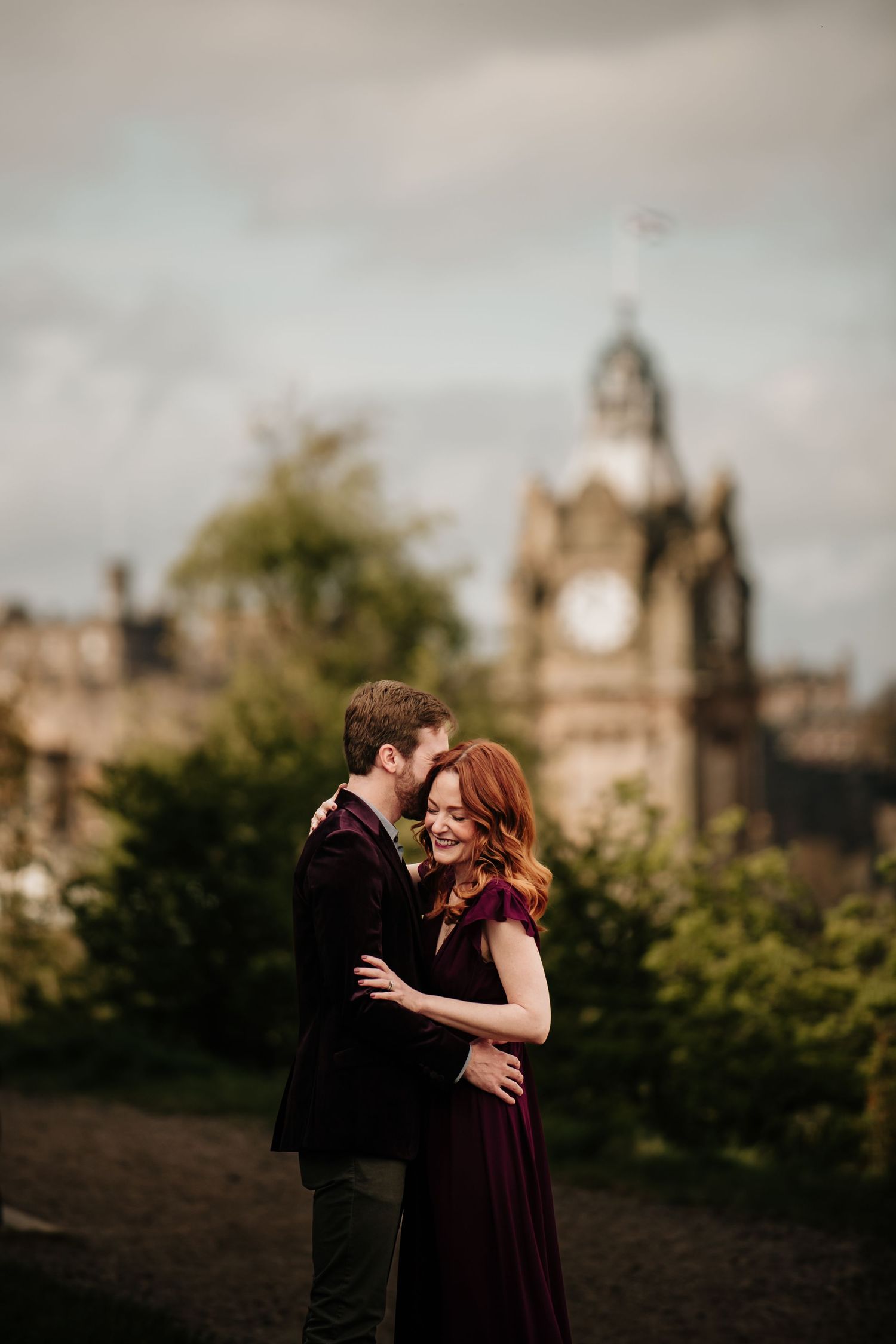 A couple embraces on Calton Hill with Edinburgh's iconic Balmoral Hotel clock tower in the background.