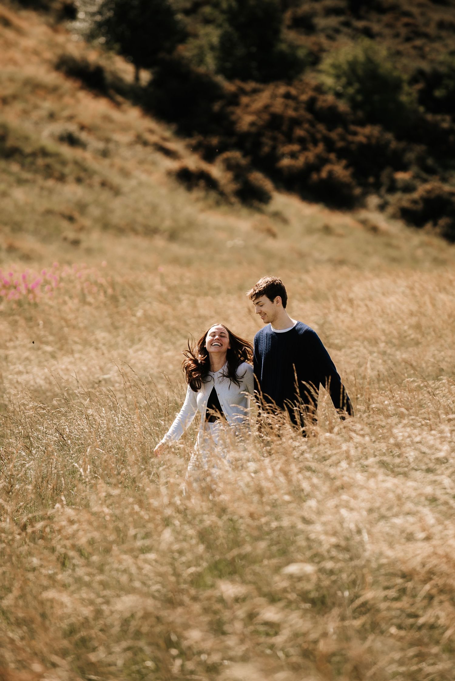 A couple walks through golden grass on a hillside during a sunny afternoon.